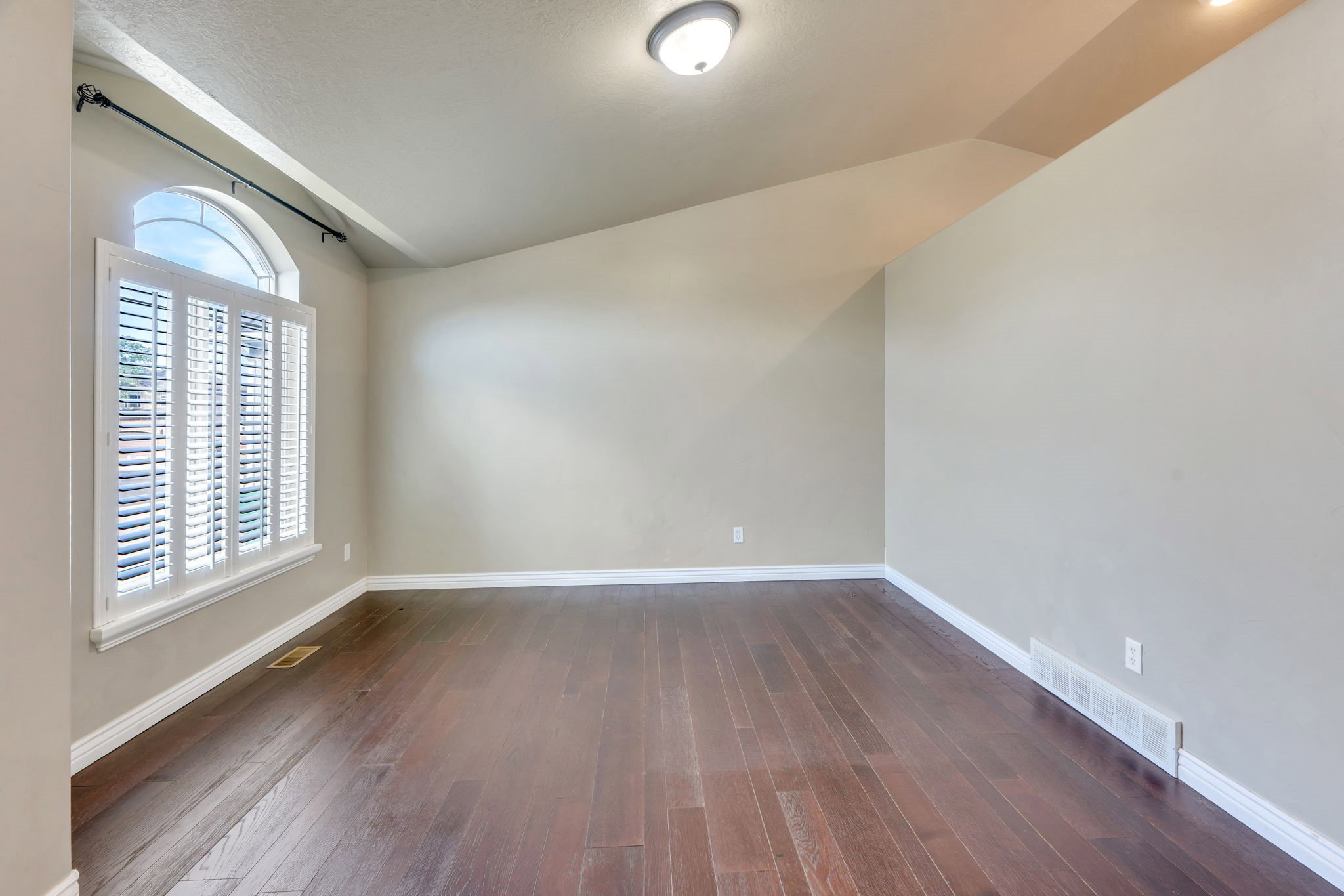 Spacious room featuring wood-finish flooring, an arched window with white plantation shutters, and a semi-flush mount ceiling light