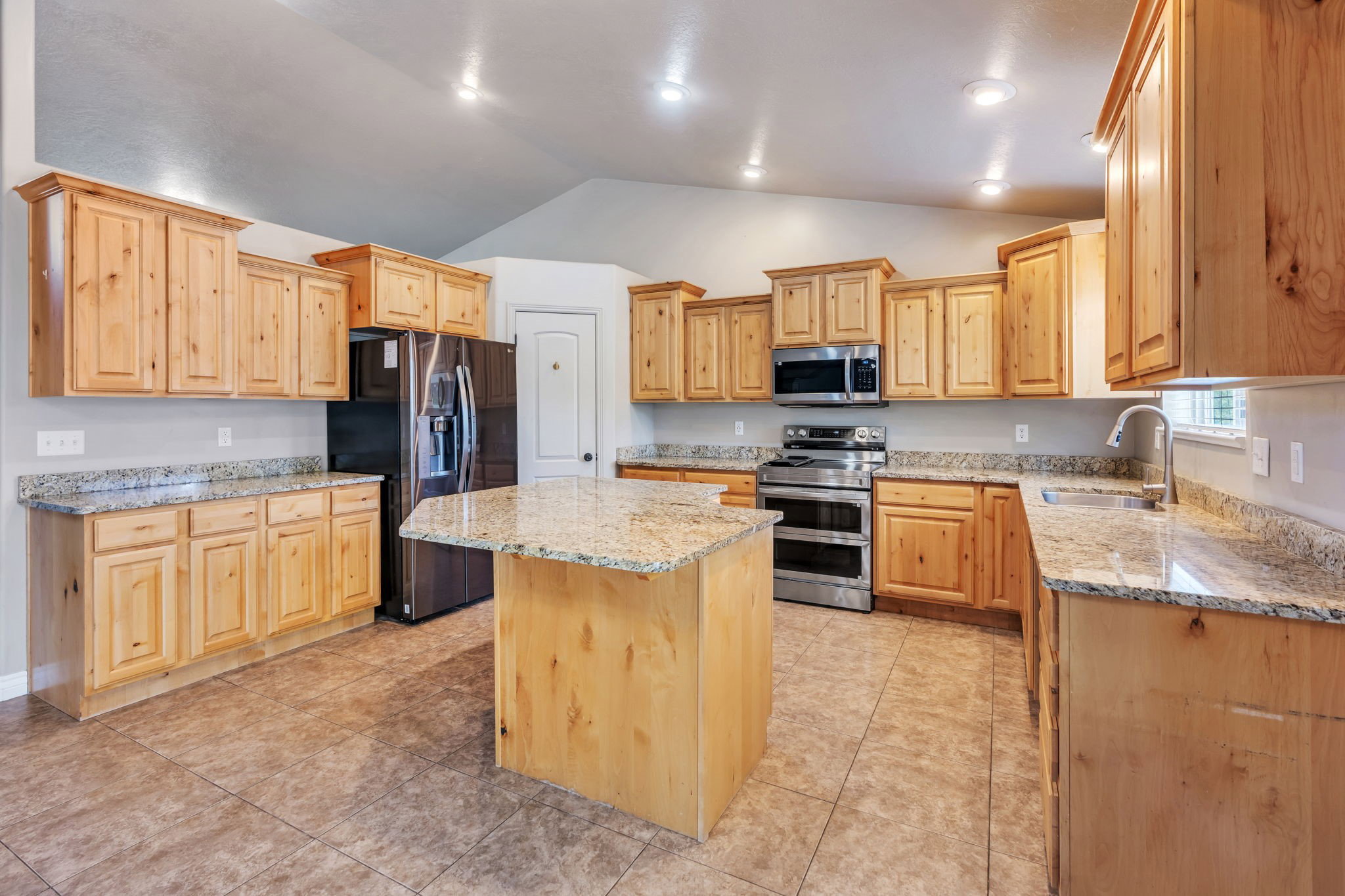 Spacious kitchen featuring an island, light wood cabinetry, stone countertops, stainless steel appliances, and tile flooring