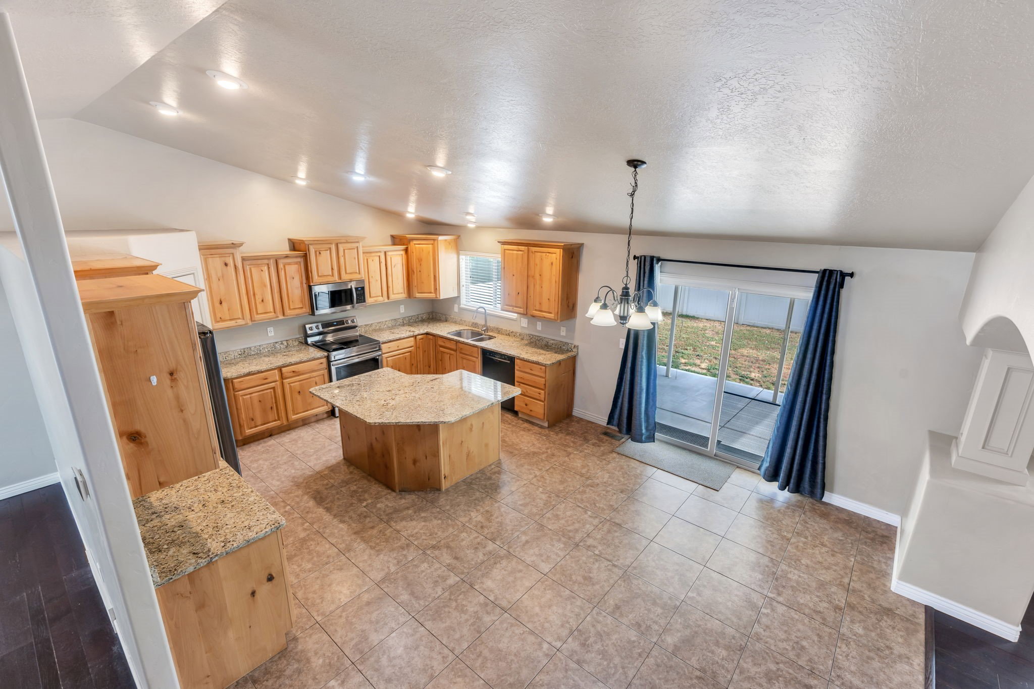 Open-concept kitchen featuring wood cabinetry, a central hexagonal island with a granite-style countertop, stainless steel appliances, and tile flooring
