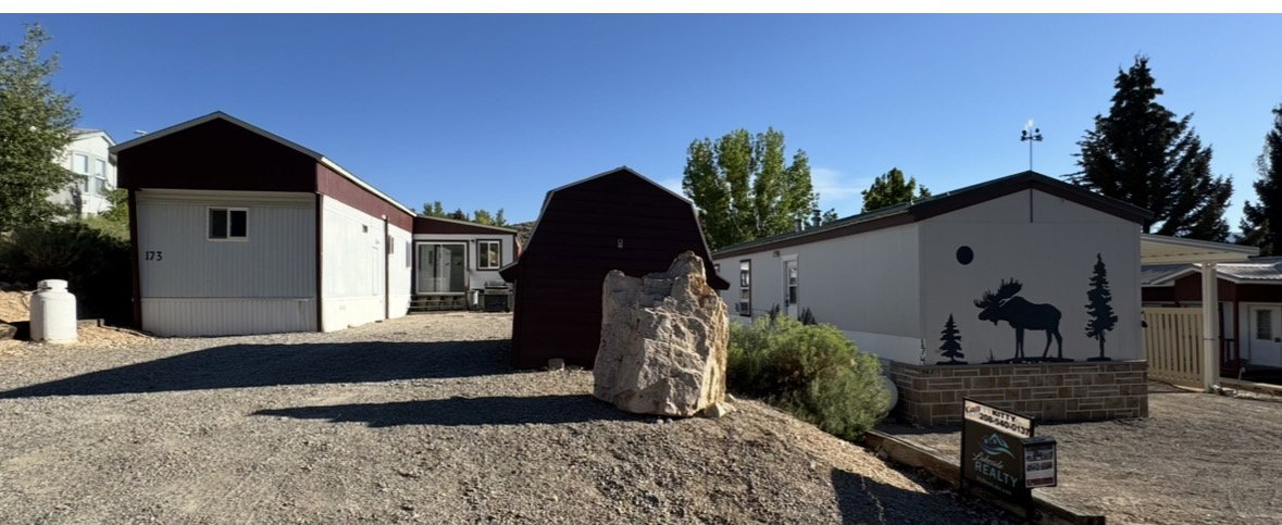 Gravel parking area fronting multiple structures, including a white-sided building with a dark roof and a separate white-sided building featuring a decorative moose and tree silhouette