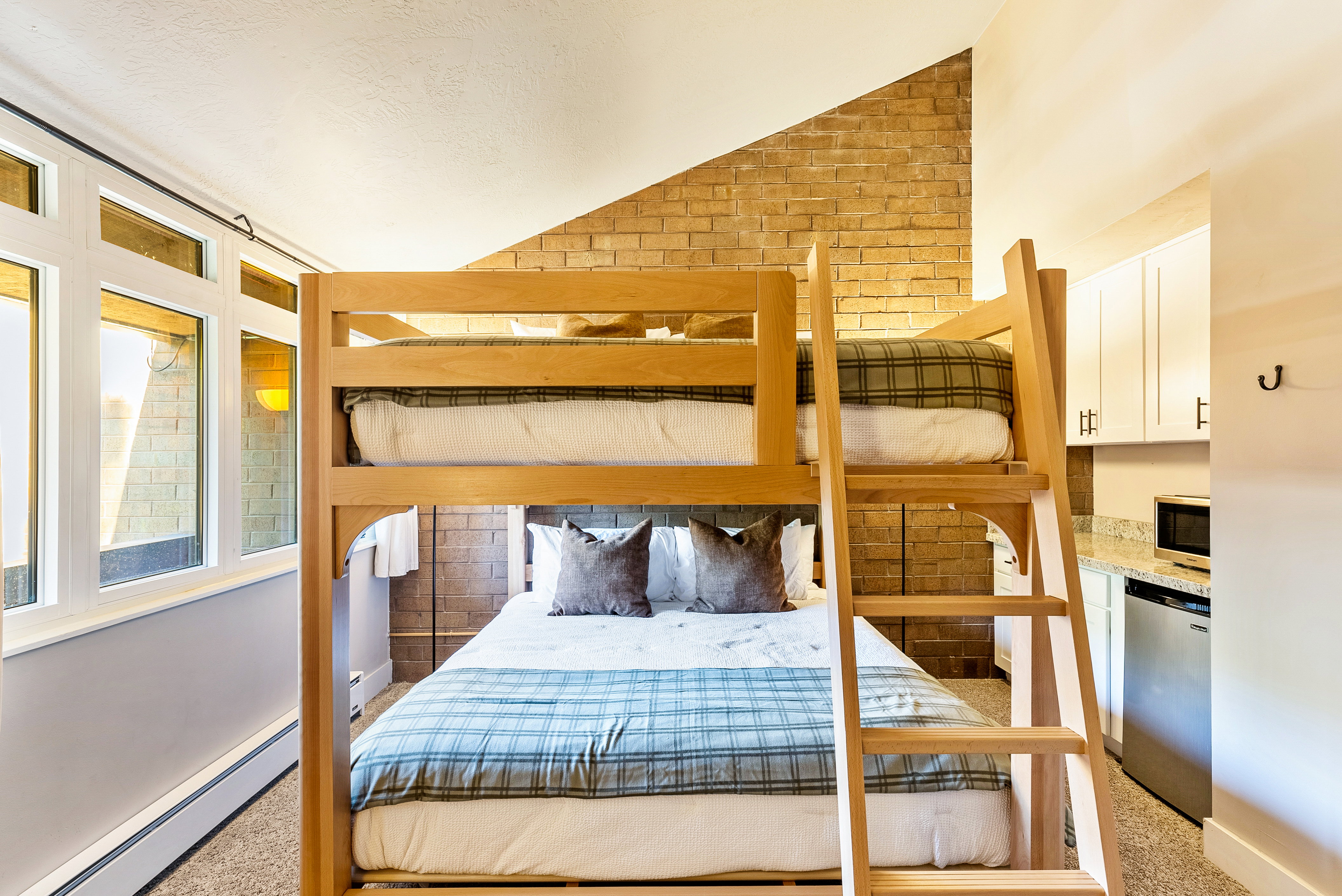 Room featuring a wood-finish bunk bed over a queen-size bed, brick accent wall, white-framed windows, and a built-in kitchenette with white cabinetry, a light-toned countertop, and a stainless steel mini-refrigerator