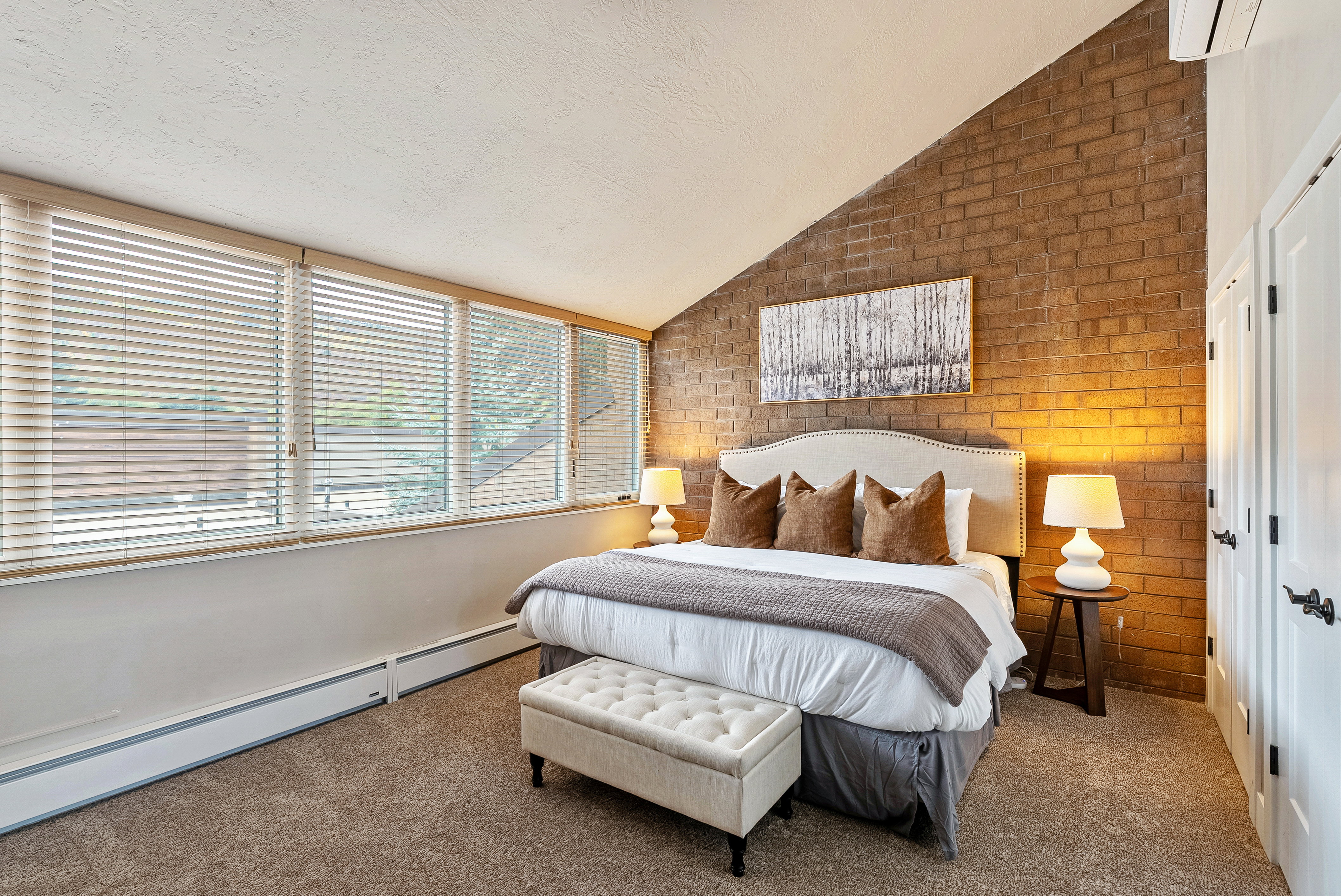Vaulted ceiling bedroom featuring an exposed brick accent wall, a series of windows with horizontal blinds, built-in white cabinetry with dark hardware, and neutral-toned carpeting