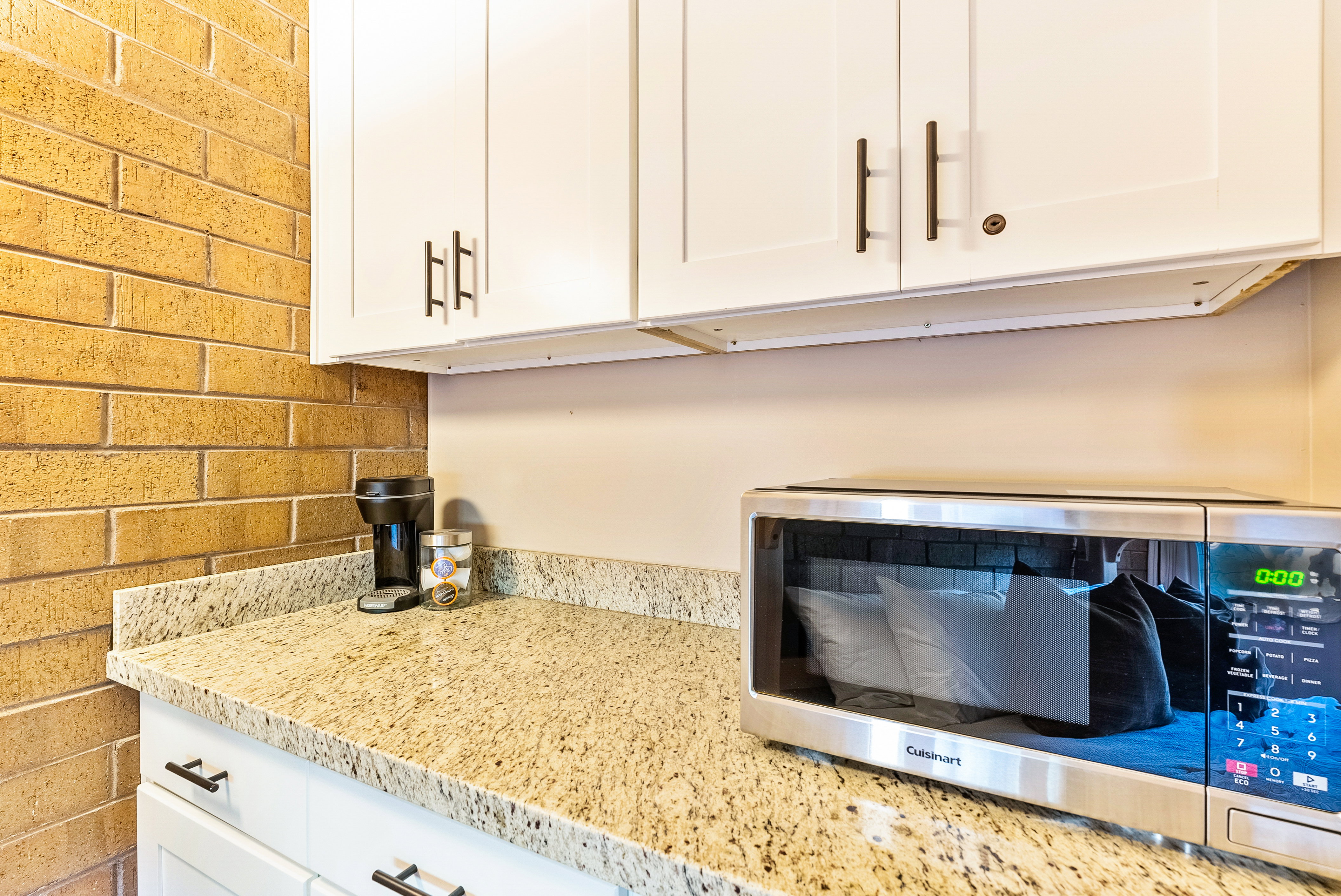 Kitchenette area featuring white shaker-style cabinetry with dark bronze hardware, a speckled granite countertop, and a brick accent wall