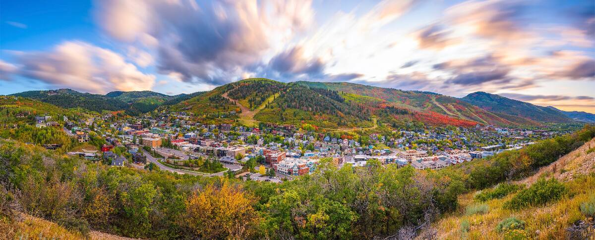 Expansive mountain landscape featuring a valley town, surrounding hillsides with natural vegetation, and a wide sky with cloud formations