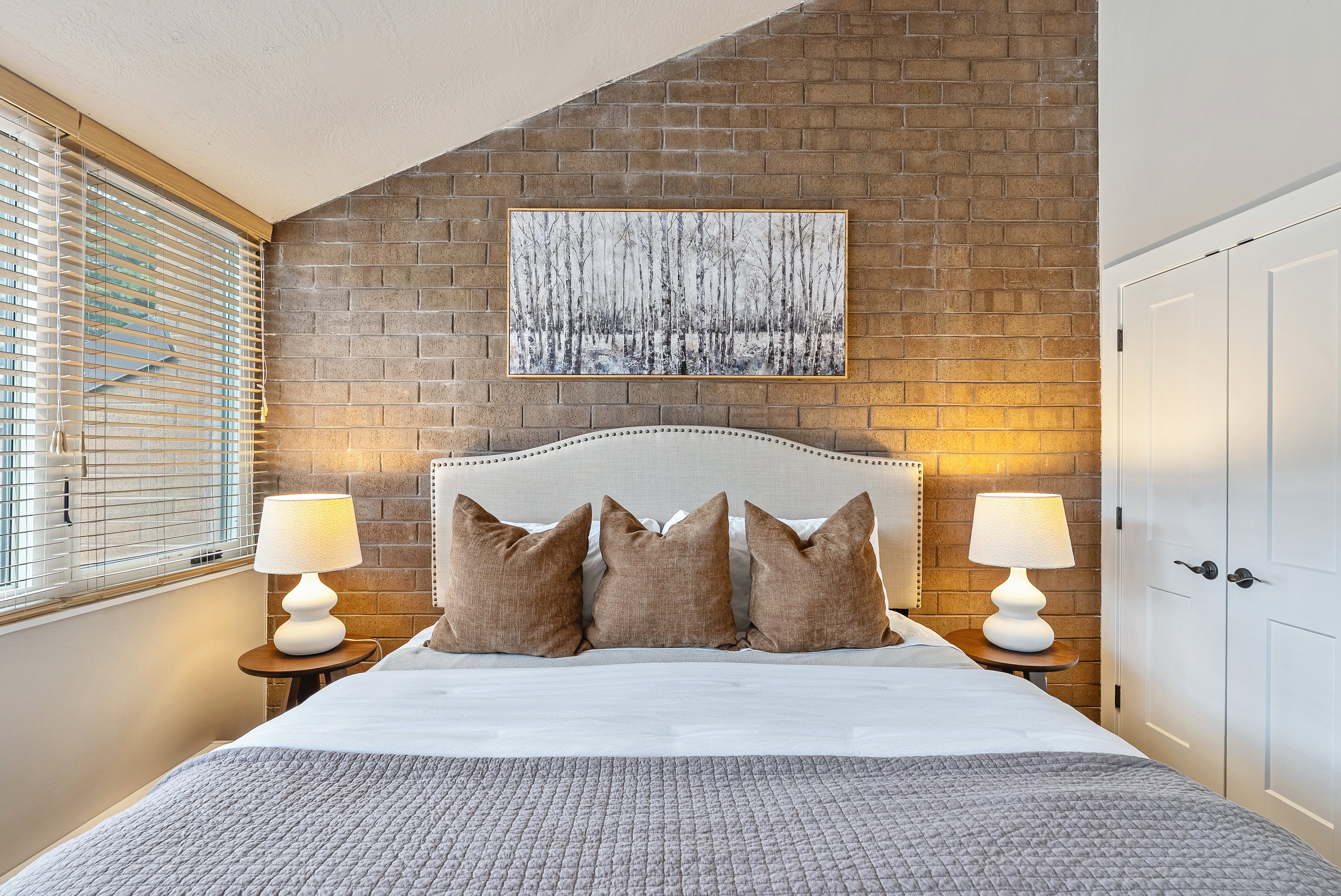 Bedroom featuring a striking exposed brick accent wall, white-paneled closet doors, and wood-finish window blinds