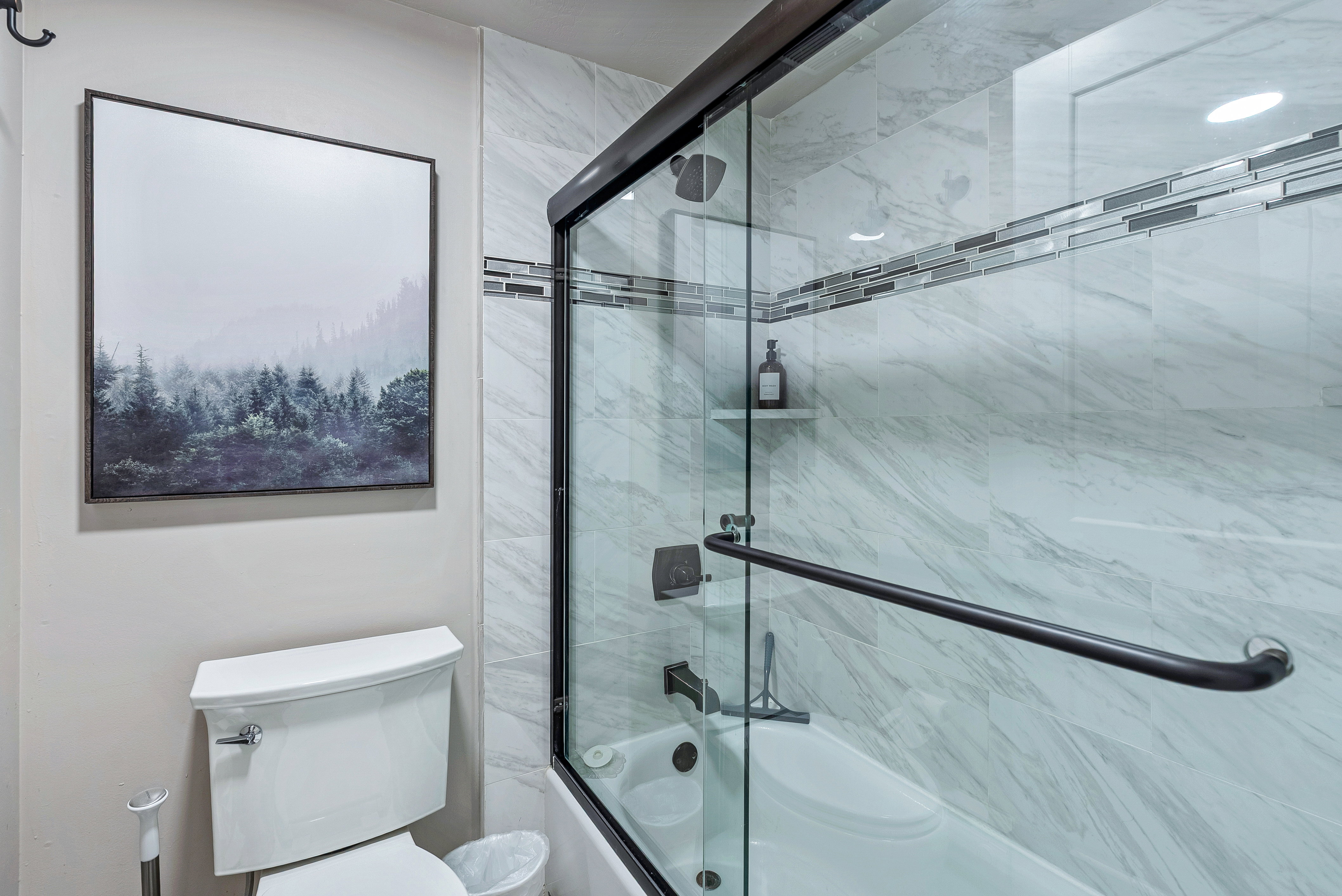 Bathtub with glass enclosure and black hardware, featuring white marble-look wall tiling and a decorative mosaic accent band
