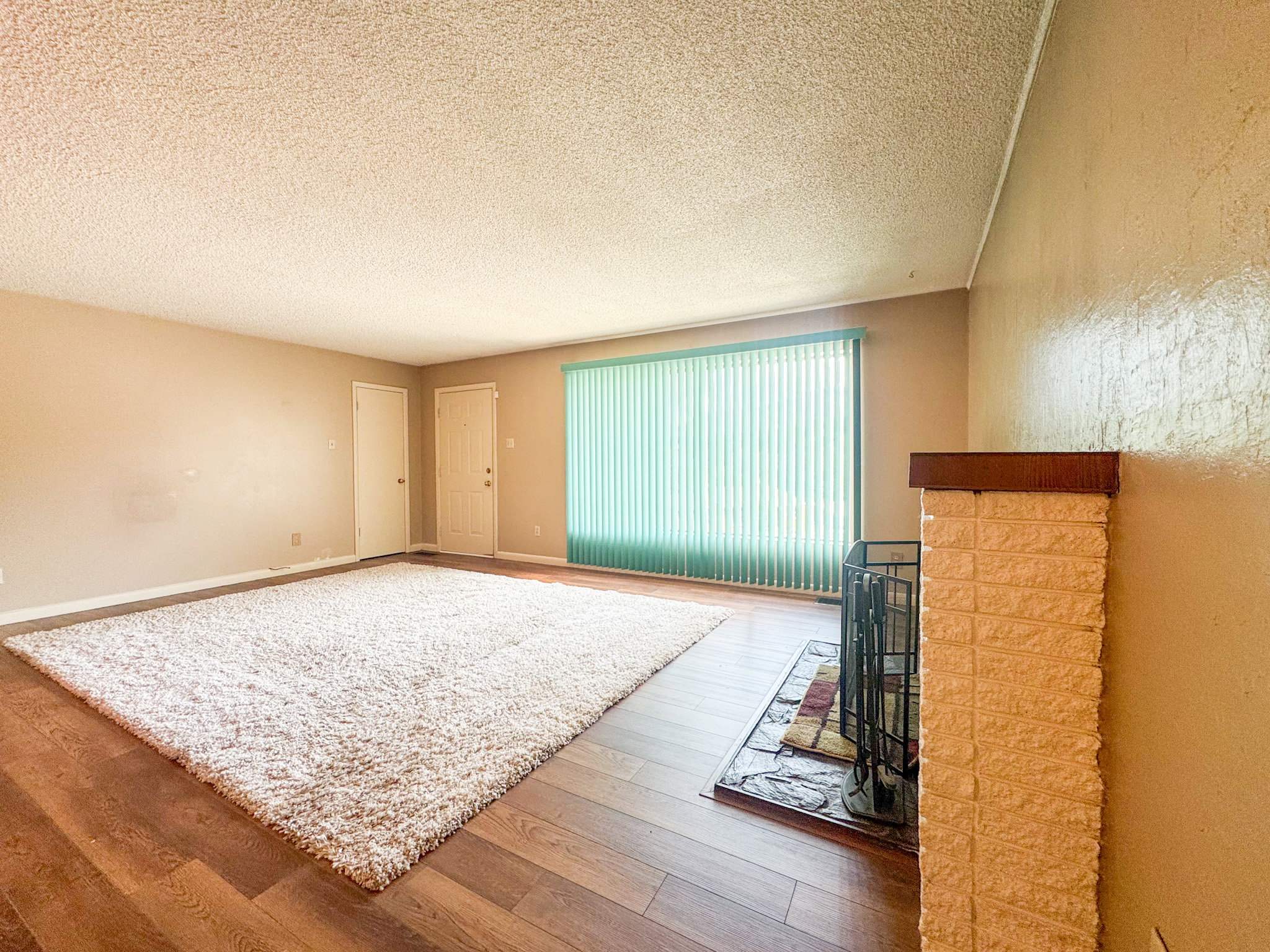 Spacious room featuring wood-finish flooring, a large window with vertical blinds, and a brick fireplace with a wood mantle