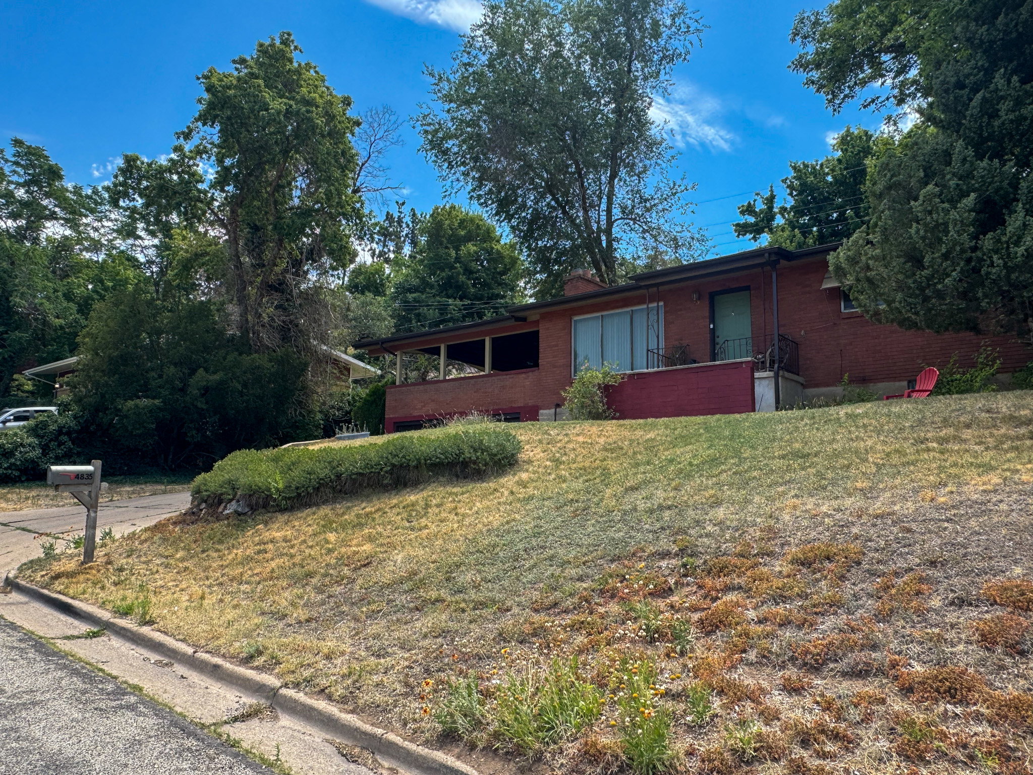 Brick facade residence featuring a covered patio, mature trees, and a sloped front yard