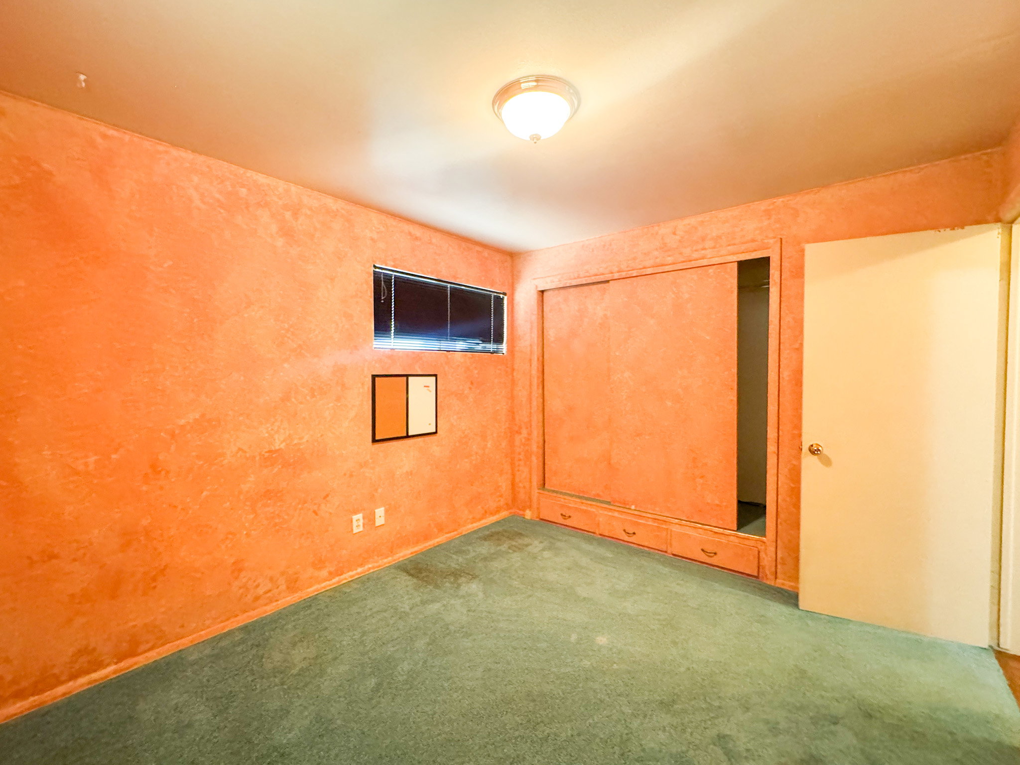 Interior room featuring textured orange walls, a horizontal window with blinds, and light green carpet flooring