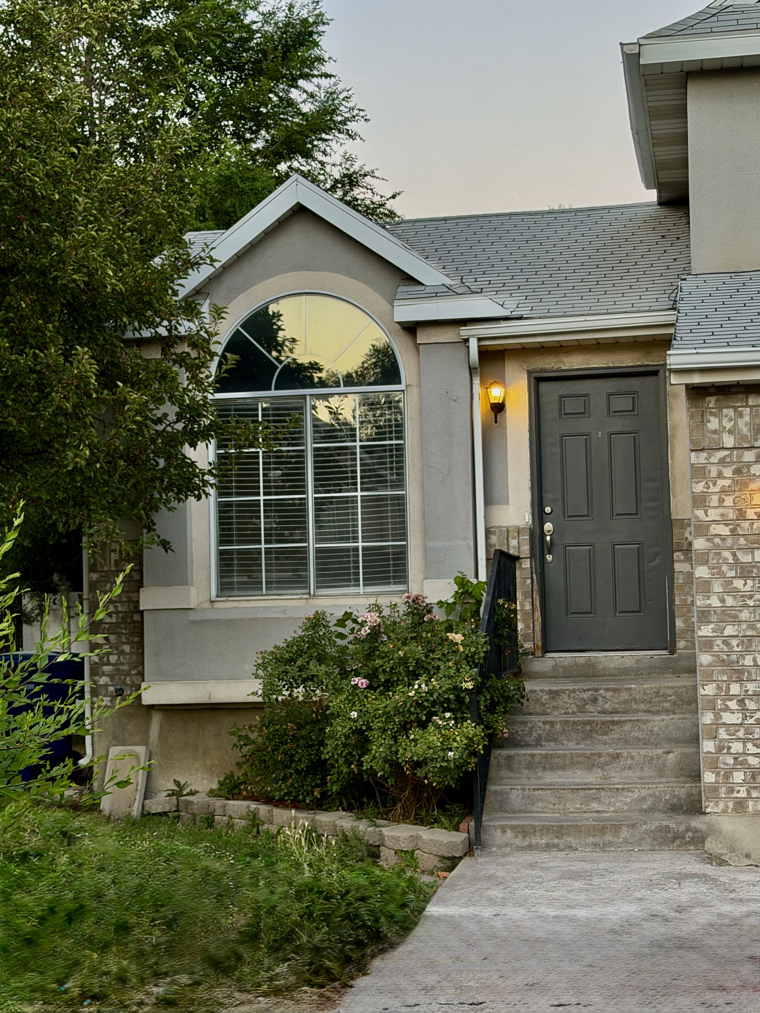 Property exterior featuring a prominent arched window, multi-pane window with horizontal blinds, concrete steps, a dark-toned entry door, and a brick facade