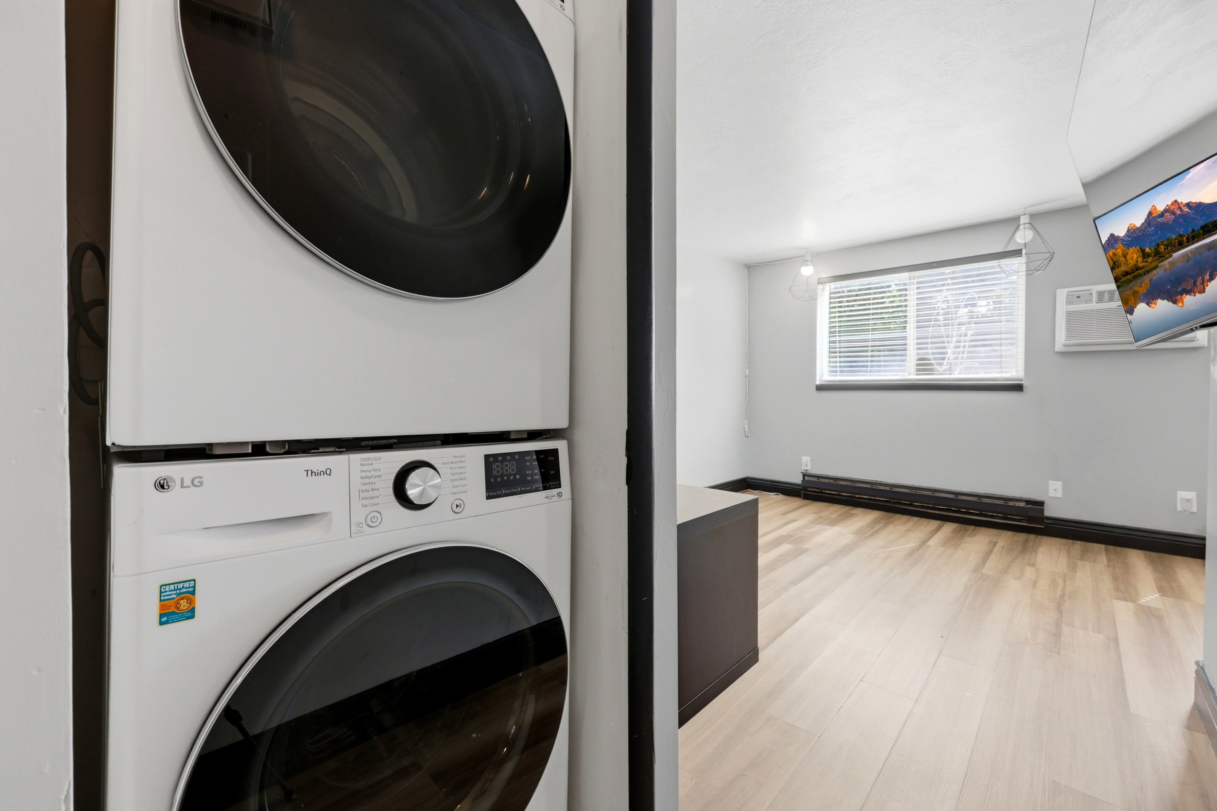 Stackable washer and dryer unit, light wood-finish flooring, recessed window with blinds, baseboard heating, and an installed wall-mounted air conditioning unit