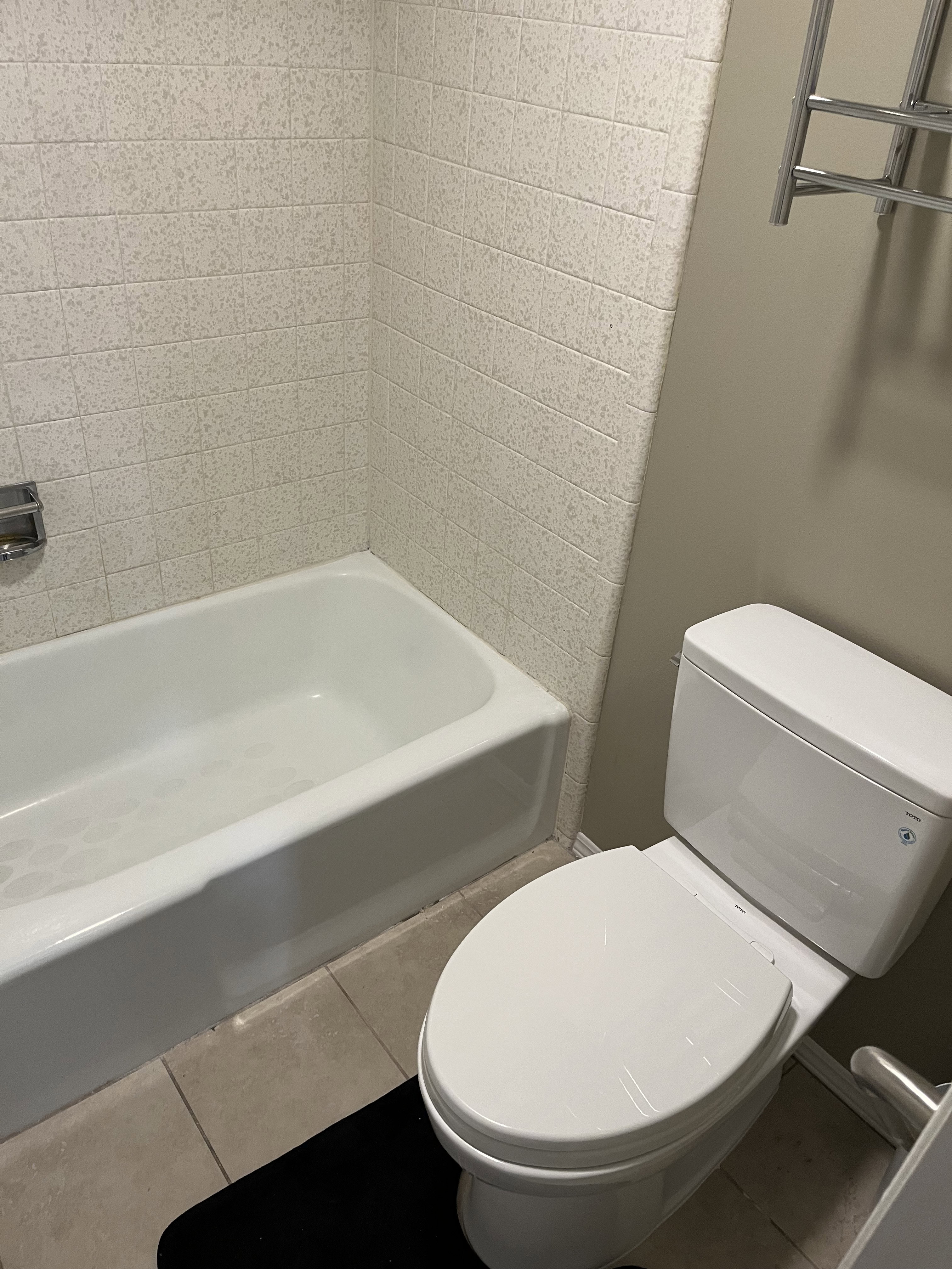 White bathtub with textured wall tiling, a modern toilet, and light-toned floor tiles