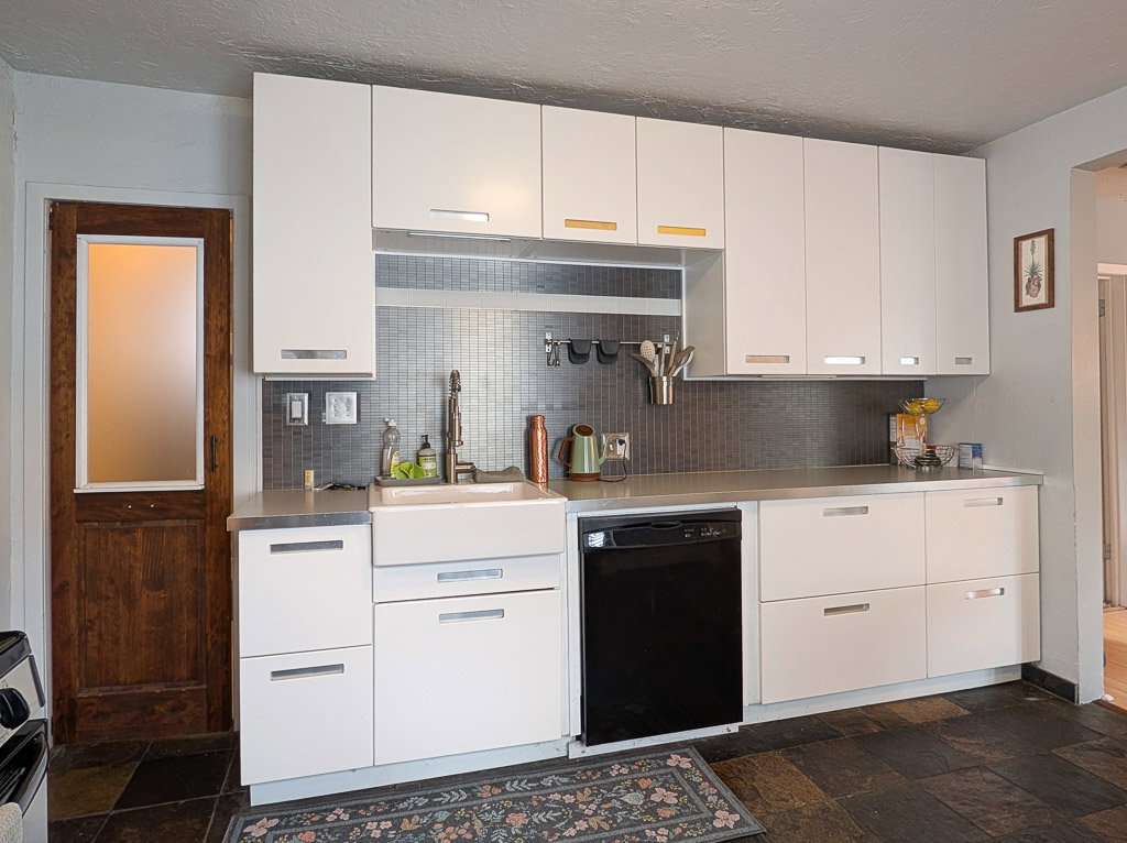 Kitchen featuring white flat-panel cabinetry, a stainless steel countertop, a mosaic tile backsplash, an integrated farmhouse sink, and slate-finish flooring