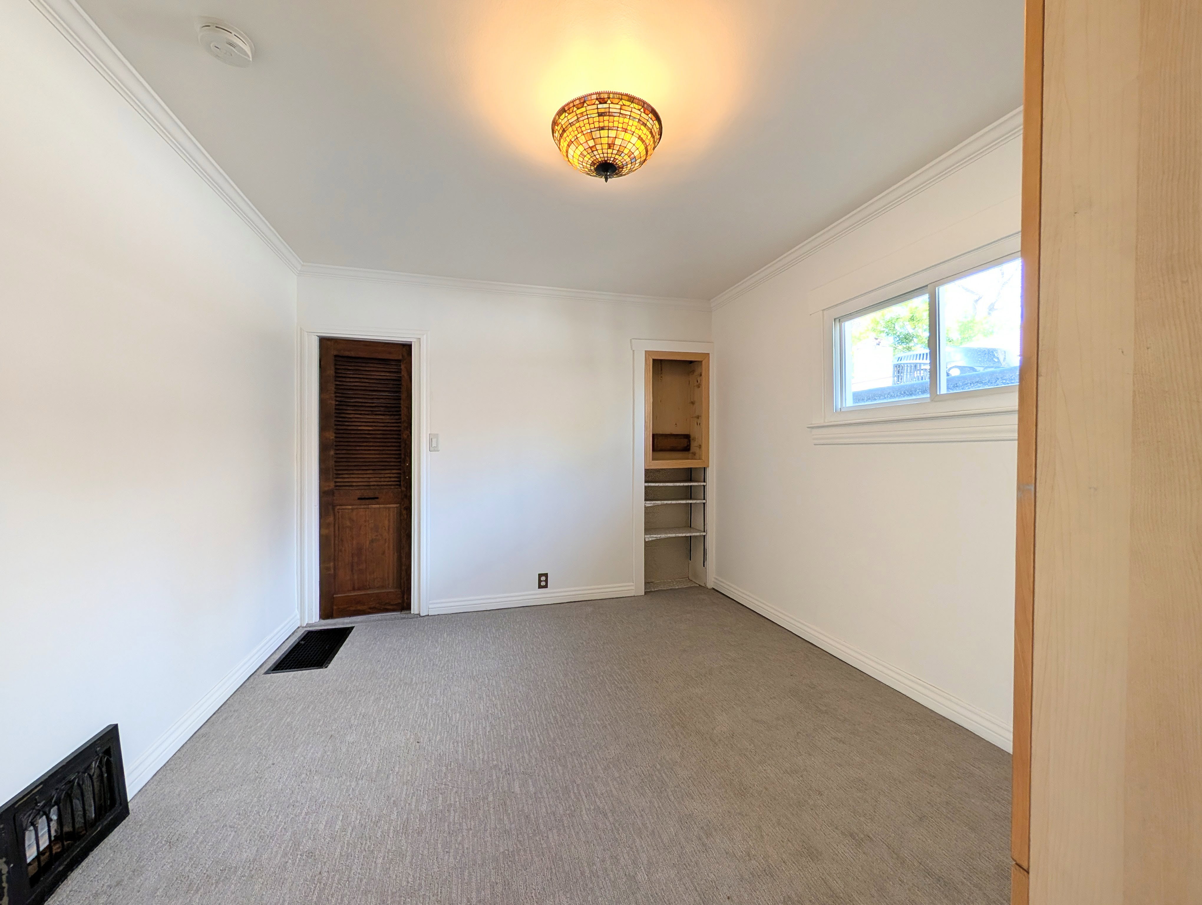 Carpeted room featuring a single window, built-in shelving, a wood louvered door, crown molding, and a decorative ceiling light fixture