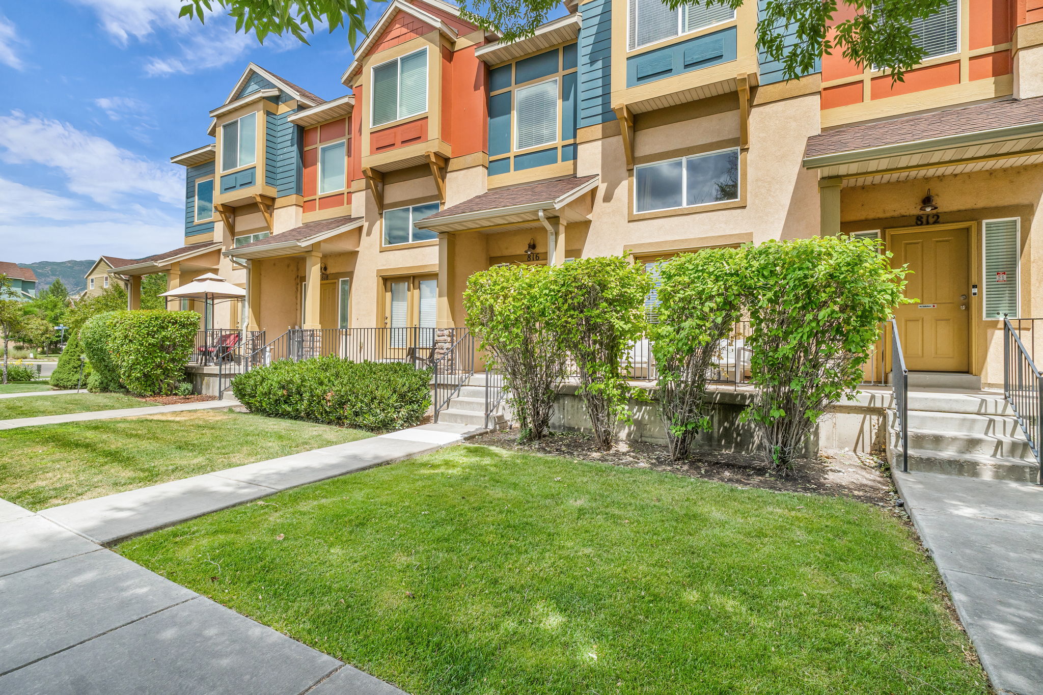 Townhome exterior featuring varied siding colors, bay windows, mature landscaping, concrete walkways, and an inviting front entrance