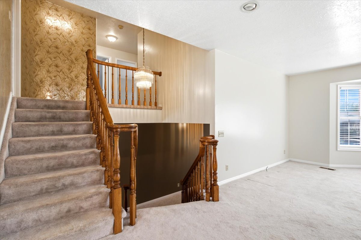 Carpeted staircase with wooden banister, featuring decorative wallpaper and a drop chandelier