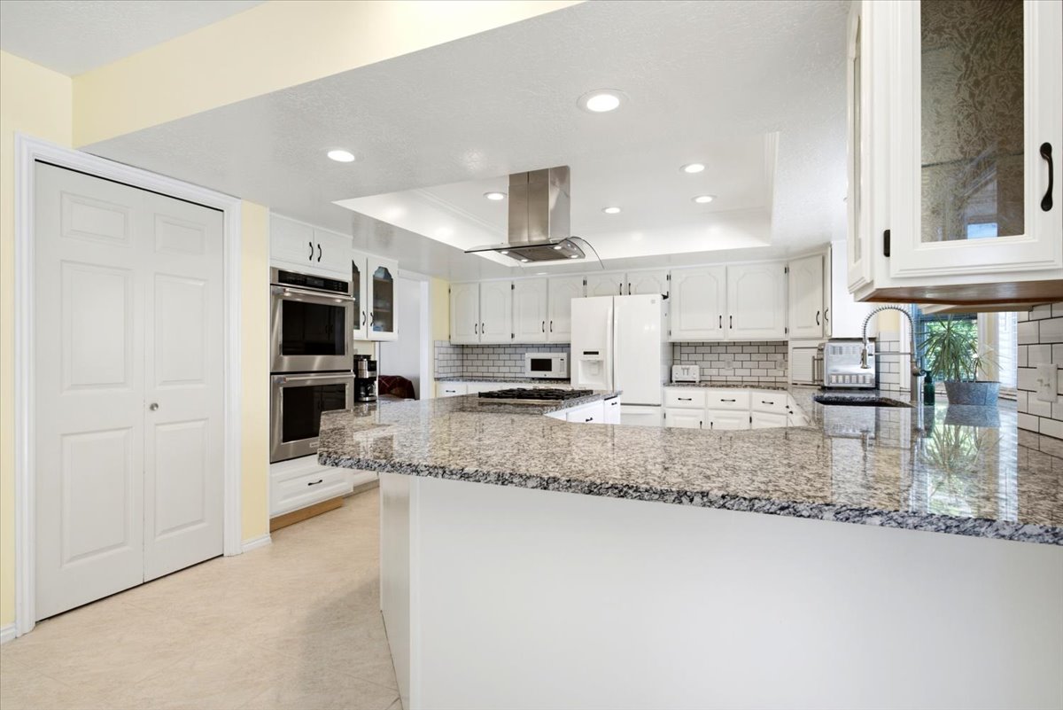 Kitchen featuring granite countertops, white cabinetry, subway tile backsplash, stainless steel range hood, and double wall ovens
