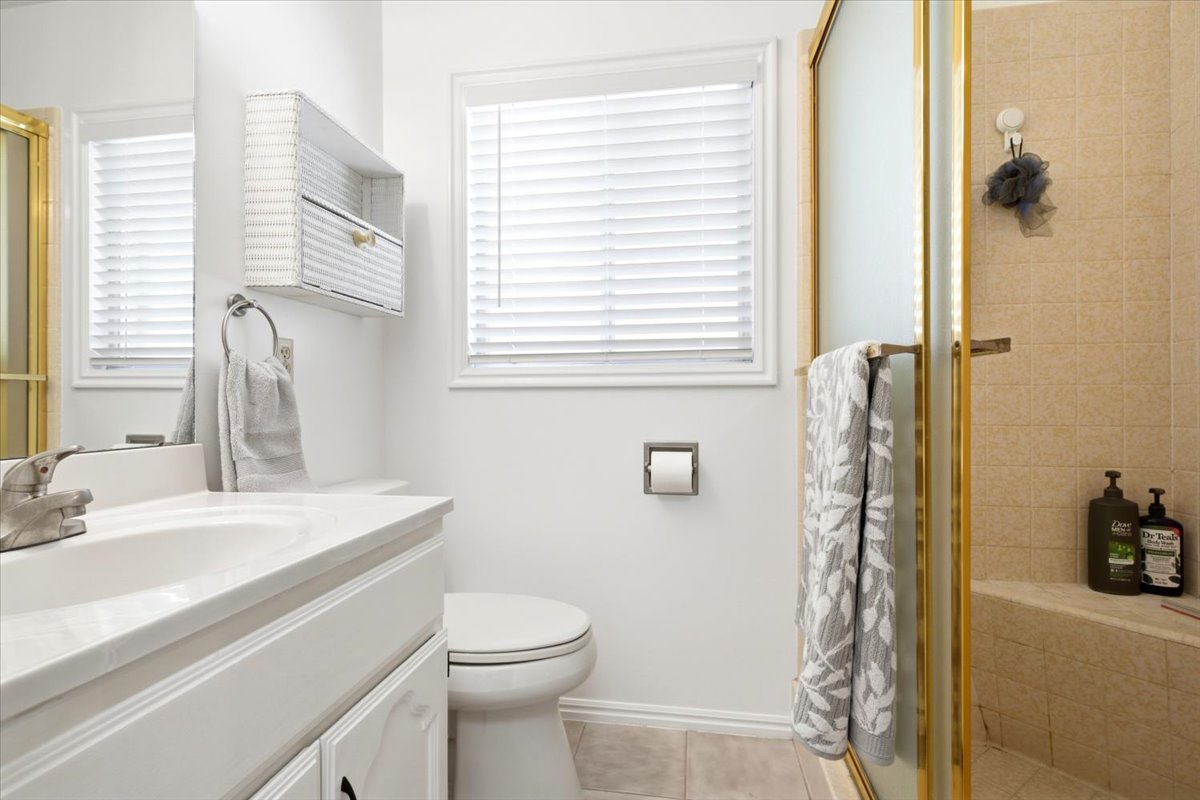 Bathroom featuring a white vanity with integrated sink, gold-framed shower enclosure, tiled shower surround with a built-in bench, and a window with blinds