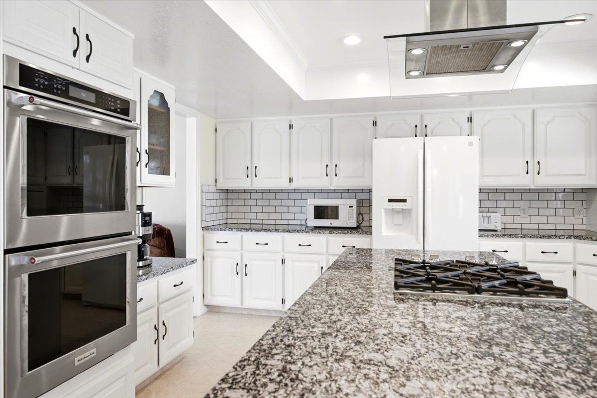 Kitchen featuring white cabinetry with black hardware, subway tile backsplash, stainless steel double ovens, and a center island with a gas cooktop and speckled countertop