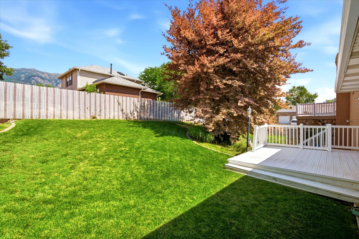 Expansive green lawn featuring a white composite deck with matching railings, a mature deciduous tree, and mountain scenery in the background