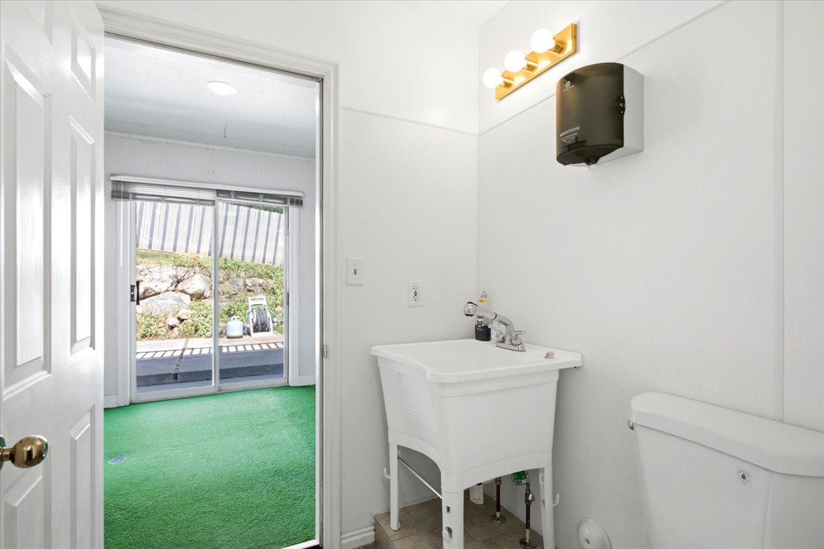 Utility room featuring a wall-mounted light fixture, service sink, toilet, and a doorway opening to a room with green flooring and sliding glass doors