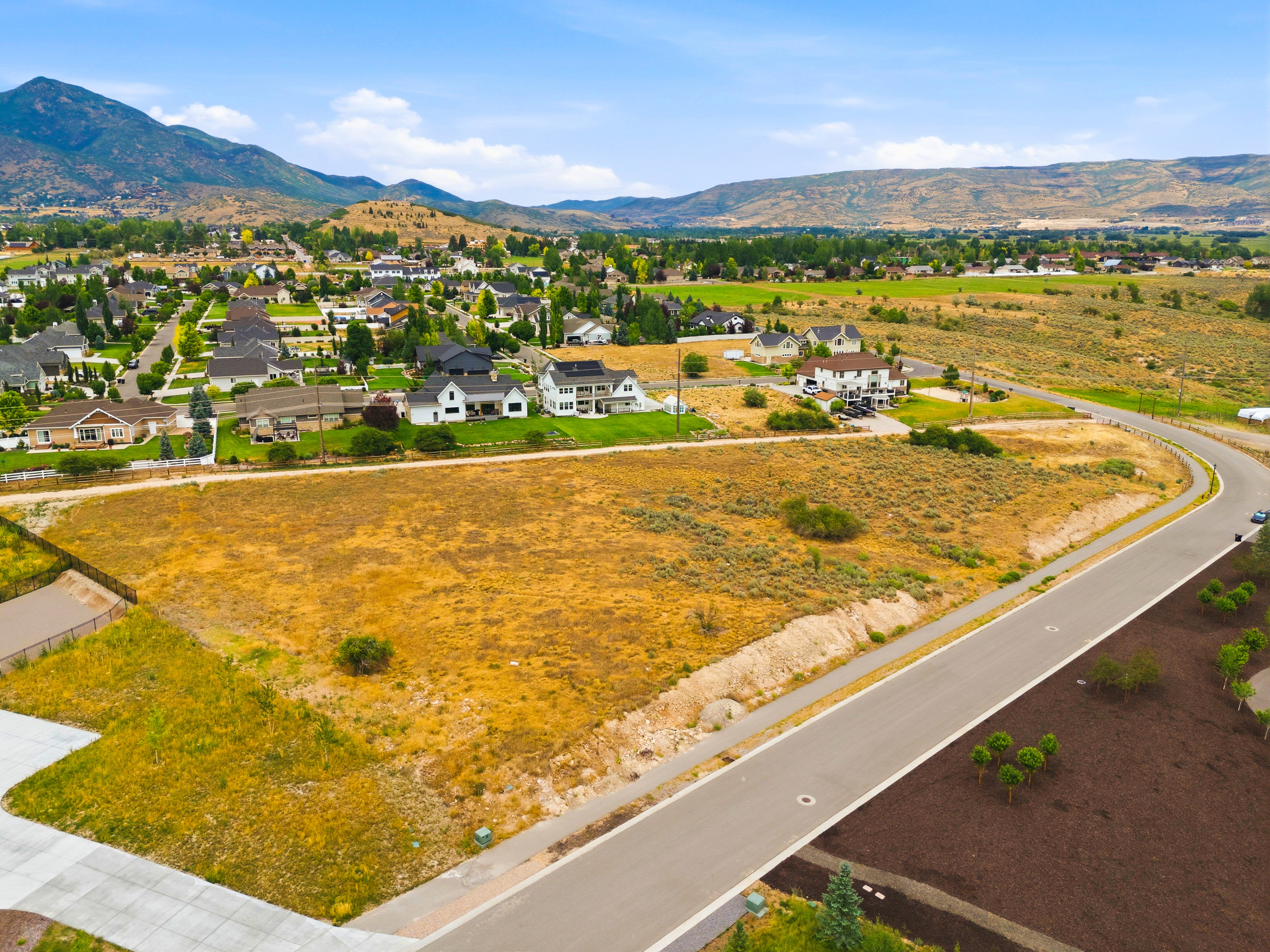 Expansive undeveloped lot presenting clear ground, paved road frontage, and established residential structures in the background