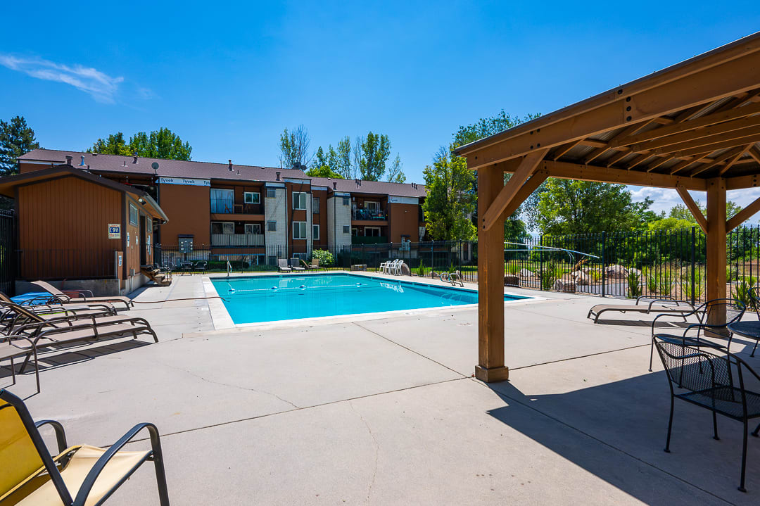 Expansive concrete pool deck featuring a rectangular swimming pool, a wooden pergola, and a brown multi-story building