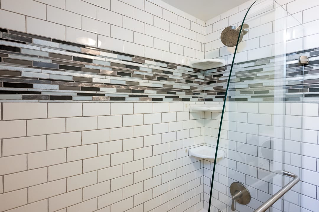 Shower enclosure featuring white subway tile, a decorative glass tile accent band, built-in corner shelves, a glass shower door, and brushed nickel fixtures