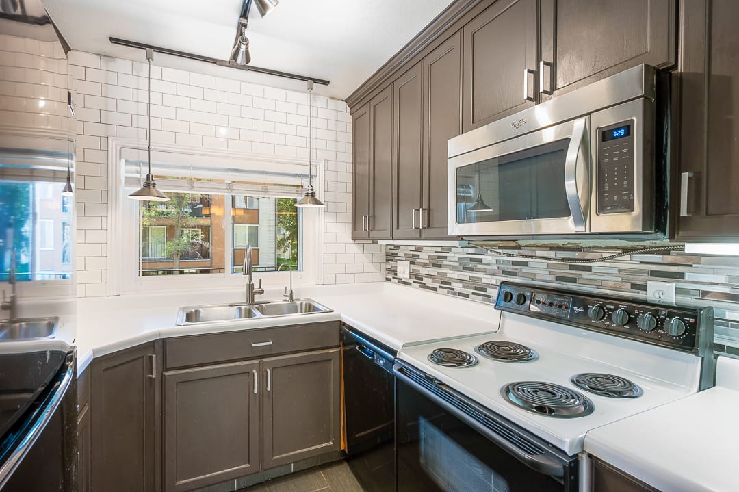 Kitchen featuring dark wood cabinetry, white countertops, a subway tile backsplash, a stainless steel microwave, and a double basin sink with a window overhead