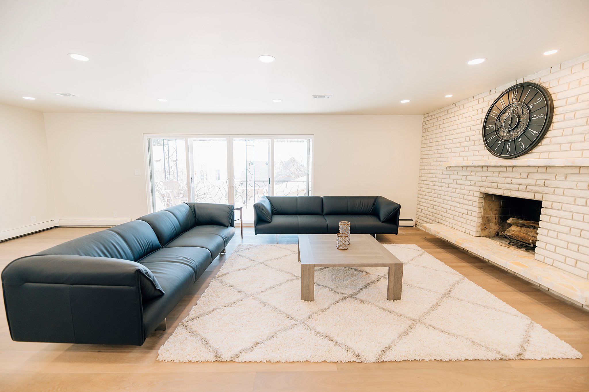 Spacious living area featuring light wood-finish flooring, a white-painted brick fireplace with a hearth, recessed ceiling lighting, and expansive glass sliding doors