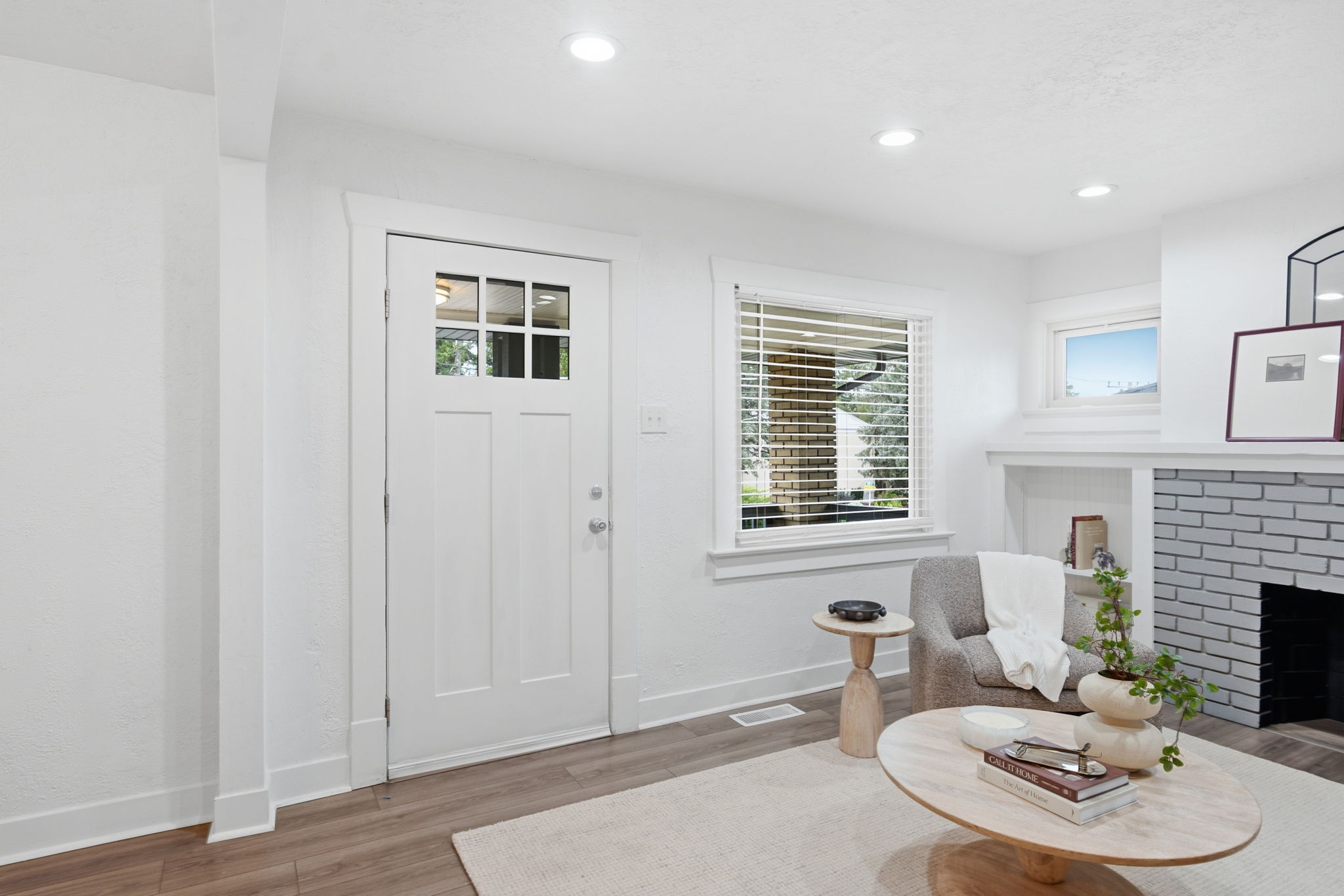 Entry door with clear glass panels, wood-finish flooring, white baseboards, recessed ceiling lighting, and a painted brick fireplace with white mantel and hearth
