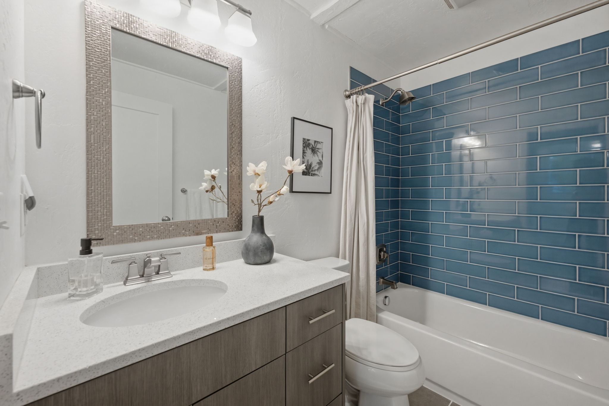 Bathroom featuring a blue subway tile bath surround, white bathtub, wood-finish vanity with an integrated sink, quartz countertop, and a decorative framed mirror