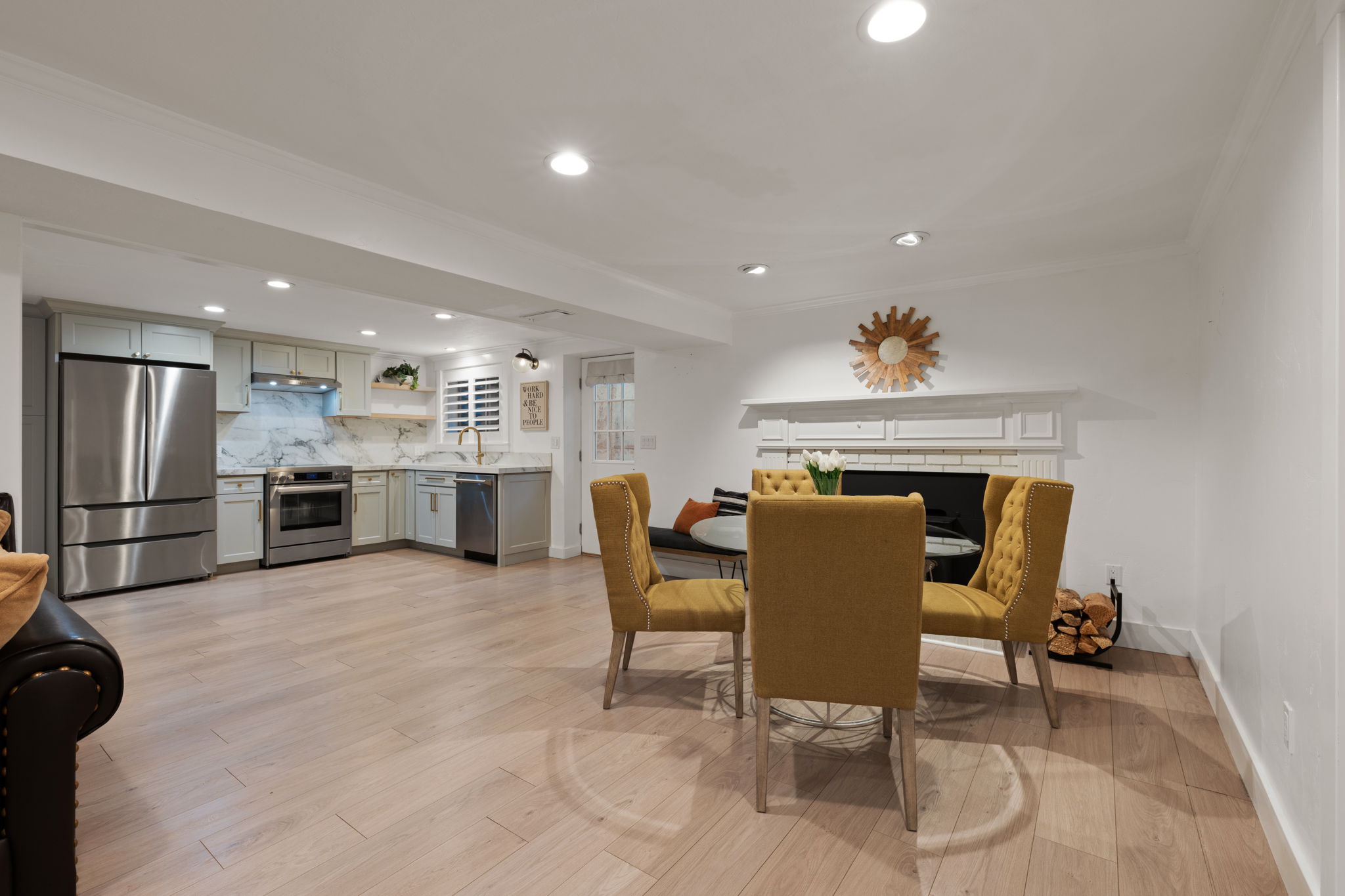 Open-concept kitchen and living area featuring light wood-finish flooring, recessed lighting, a built-in fireplace with a white mantel, stainless steel appliances, and white cabinetry with a light-colored backsplash