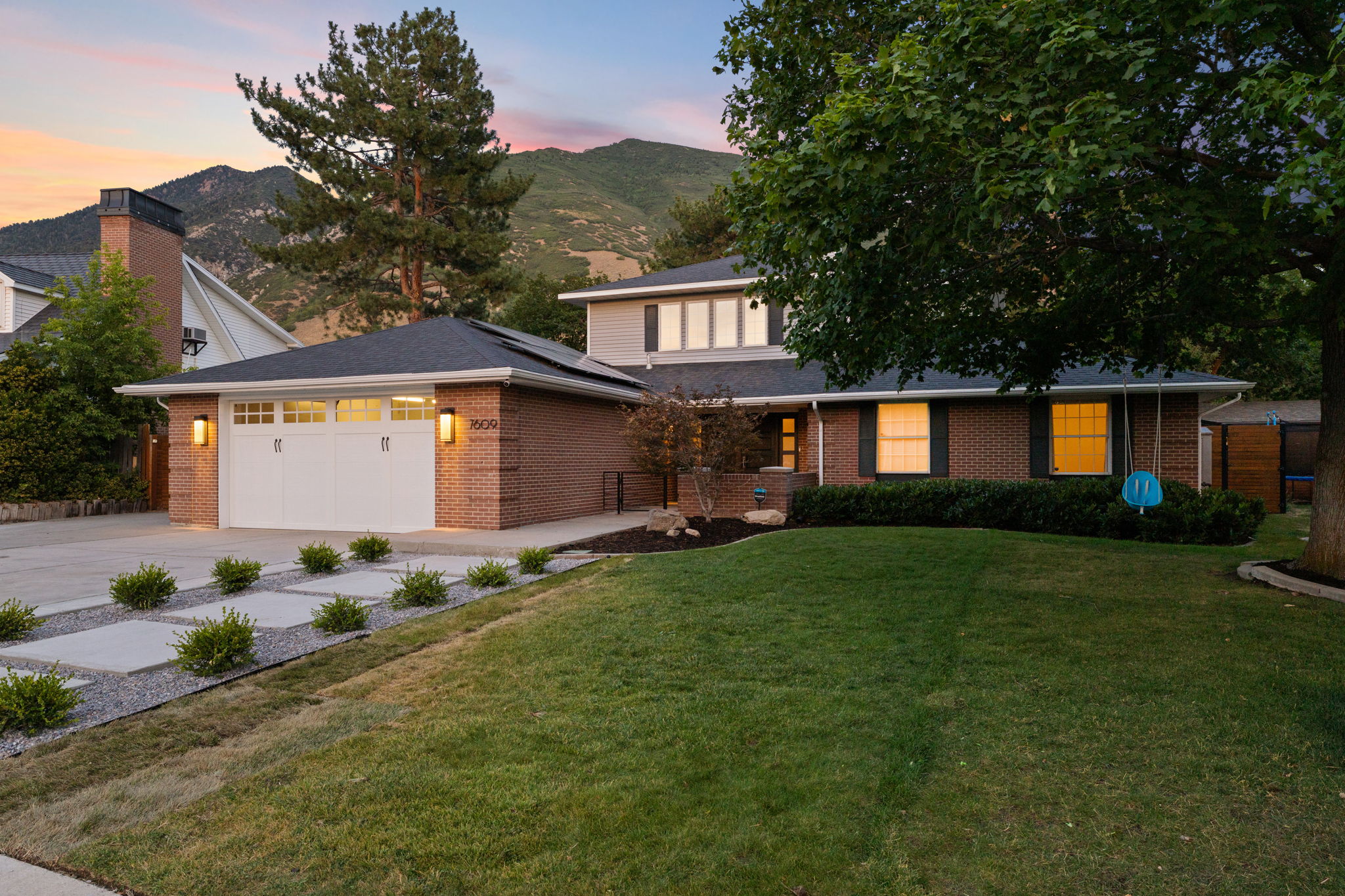 Brick facade home featuring a two-car garage with transom windows, a concrete driveway with decorative pavers, a manicured front lawn, and mature trees