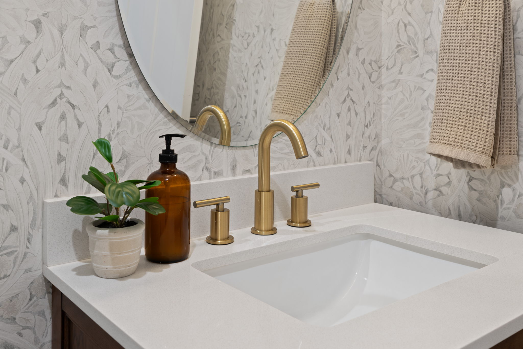 Integrated rectangular sink with a light-toned countertop, a three-piece gold-toned faucet set, a circular mirror, and patterned wallpaper