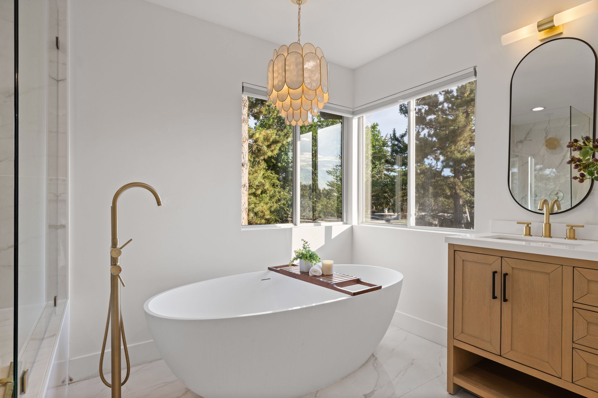 Freestanding soaking tub with a brass floor-mounted filler, corner windows, a wood-finish vanity with a white countertop, an oval mirror, and a decorative chandelier