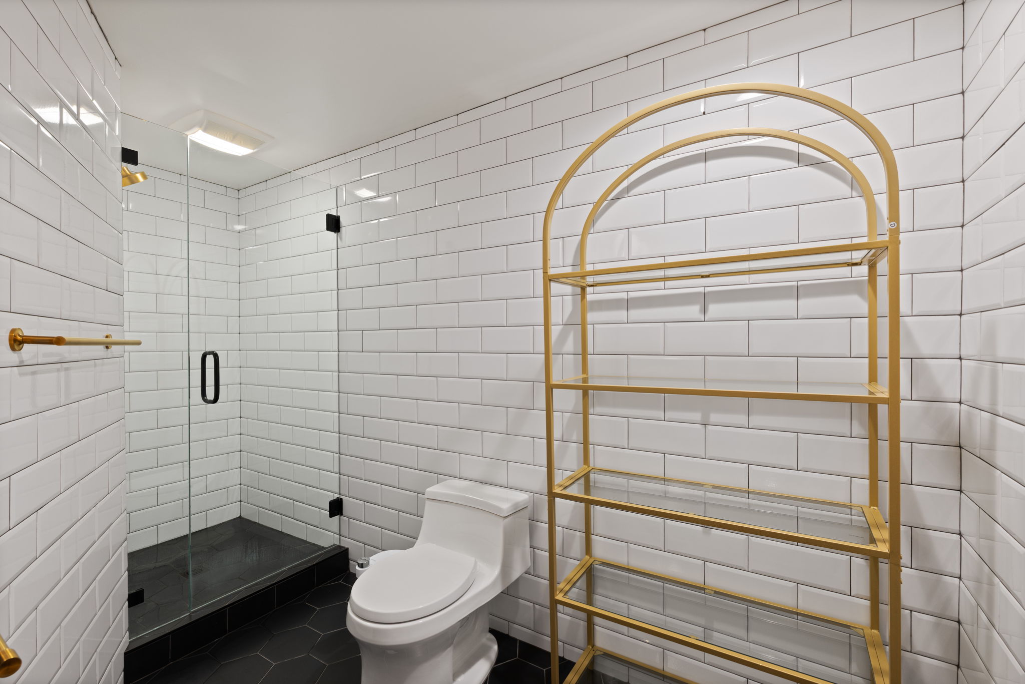 Bathroom featuring a stand-up shower with clear glass enclosure and black base, white subway tile walls, black hexagonal floor tiles, and gold-finish fixtures