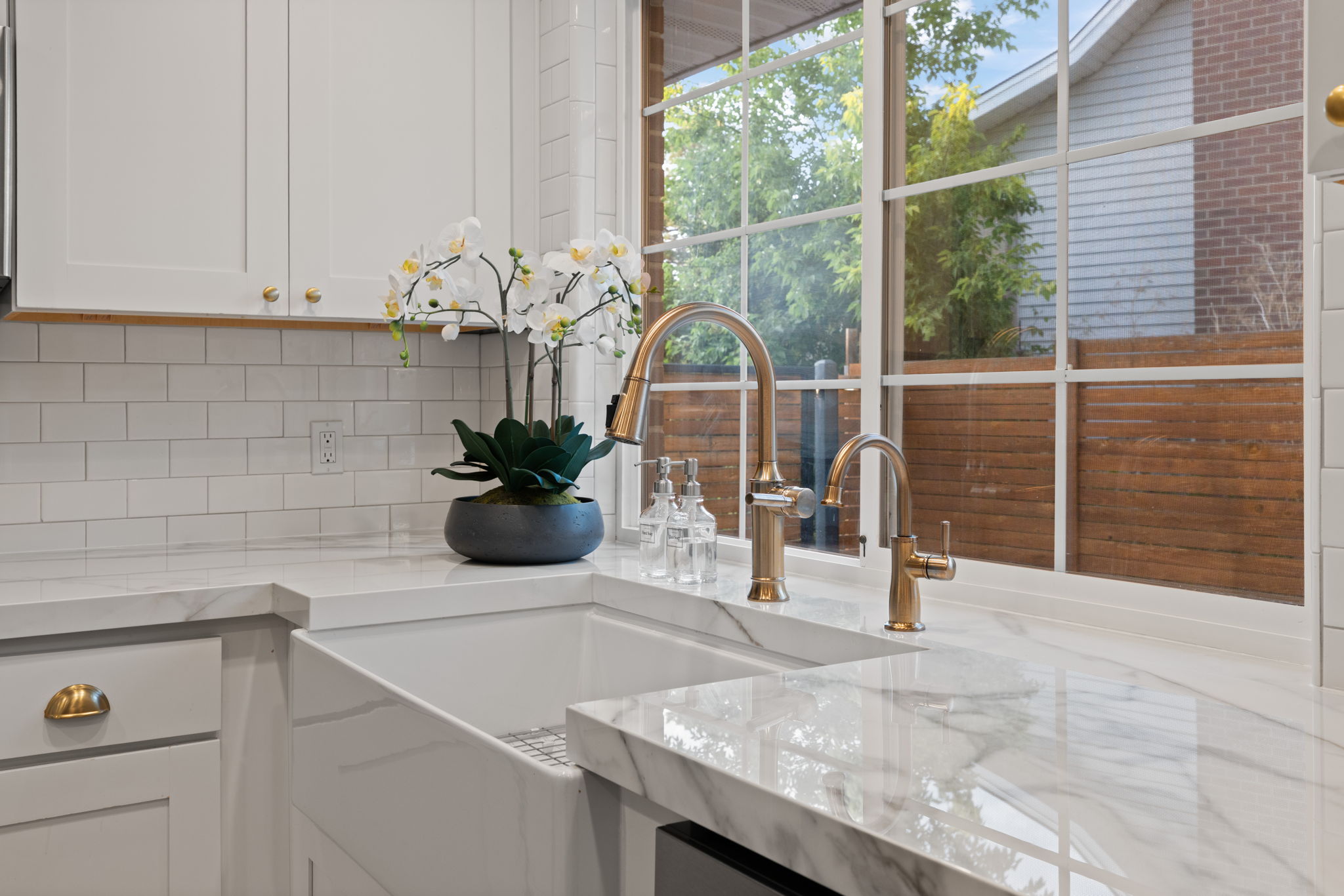 Kitchen workstation featuring a large farmhouse sink, dual brass-finish faucets, white shaker-style cabinetry, and white subway tile backsplash