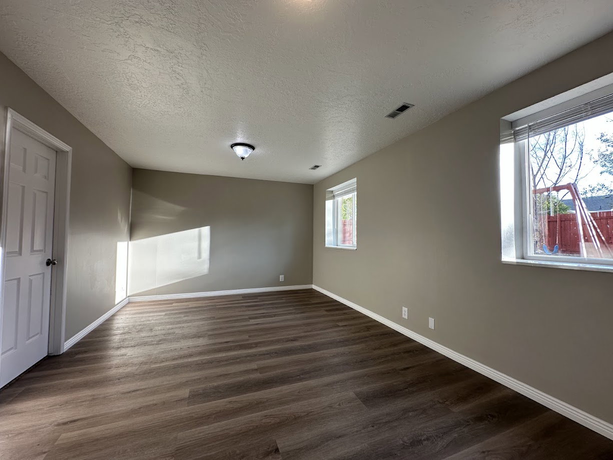 Spacious interior room featuring wood-finish flooring, two windows with blinds, a white paneled door, and neutral wall paint