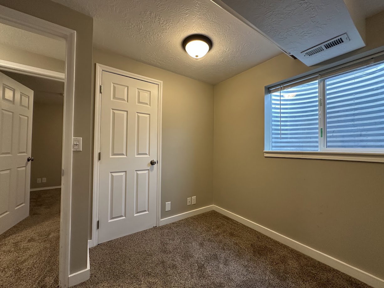 Carpeted room featuring a white paneled door, a white-framed window with blinds, a ceiling-mounted light fixture, and neutral-toned wall paint