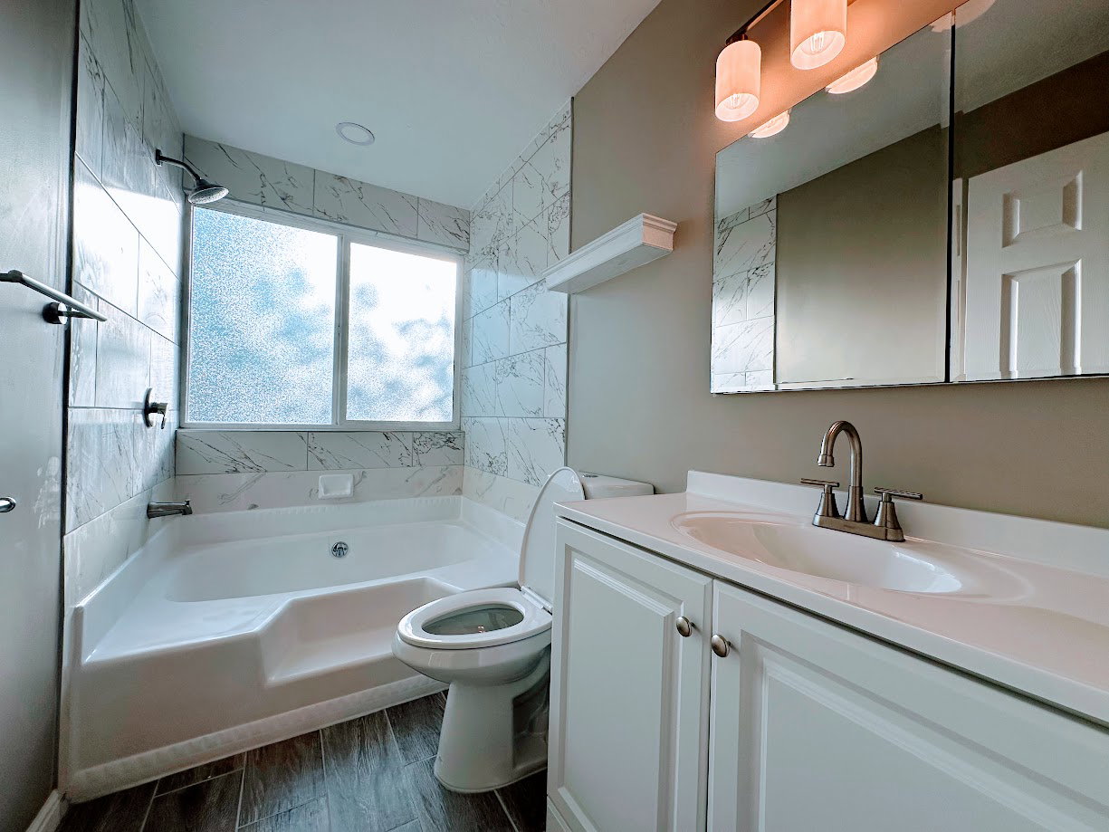 Bathroom featuring a built-in tub with shower, marble-finish tile surround, white vanity with integrated sink, contemporary vanity lighting, and wood-finish flooring