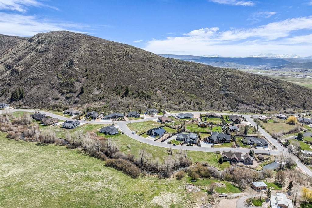 Aerial perspective showcasing a residential community nestled at the base of a verdant mountain, featuring a curving street layout and distant snow-capped peaks