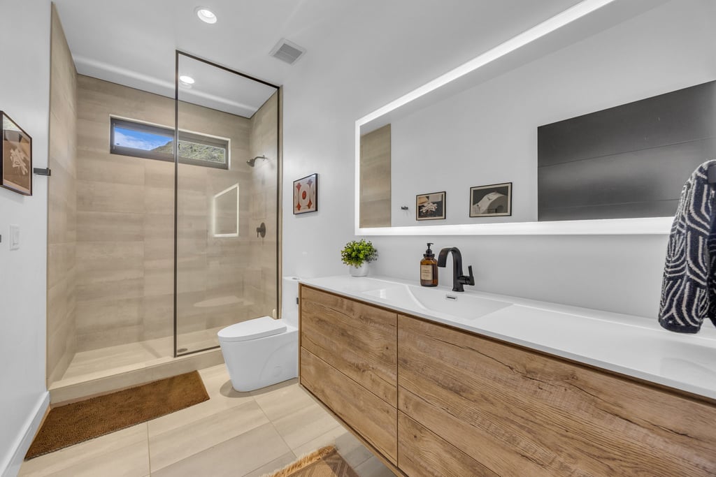 Contemporary bathroom featuring a floating wood-finish vanity with an integrated sink, a large back-lit mirror, a glass-enclosed shower, a wall-mounted showerhead, and a horizontal window