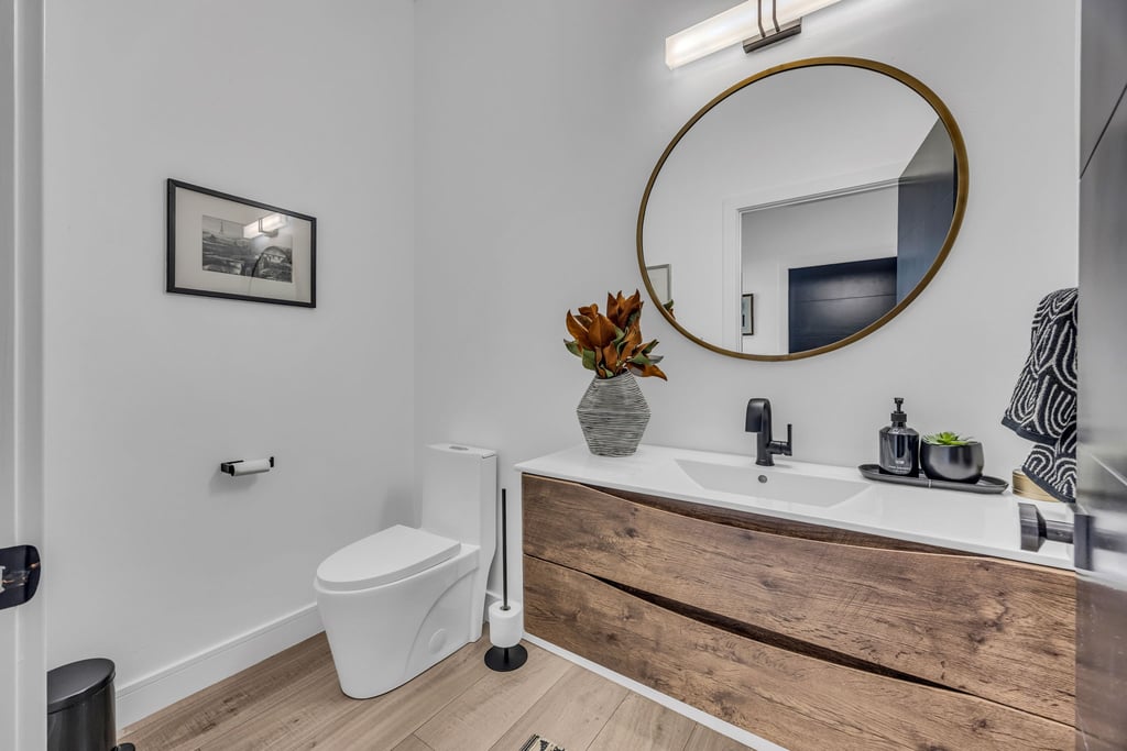 Modern bathroom featuring a wood-finish vanity with an integrated white sink and dark bronze faucet, a circular mirror with a bronze frame, and wood-finish flooring