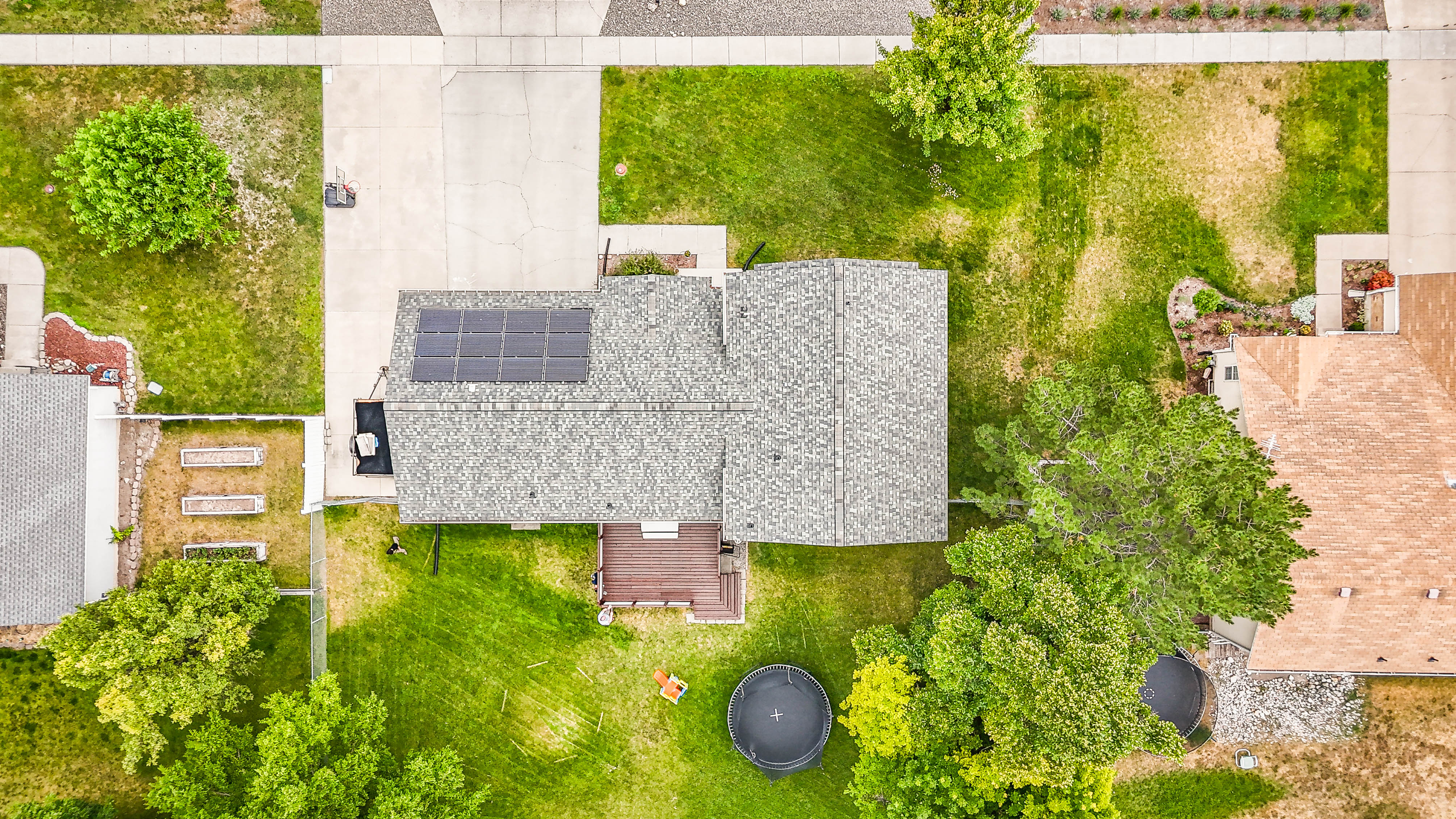 Aerial property view featuring solar panels, a gabled roof, an expansive concrete driveway, and mature landscaping