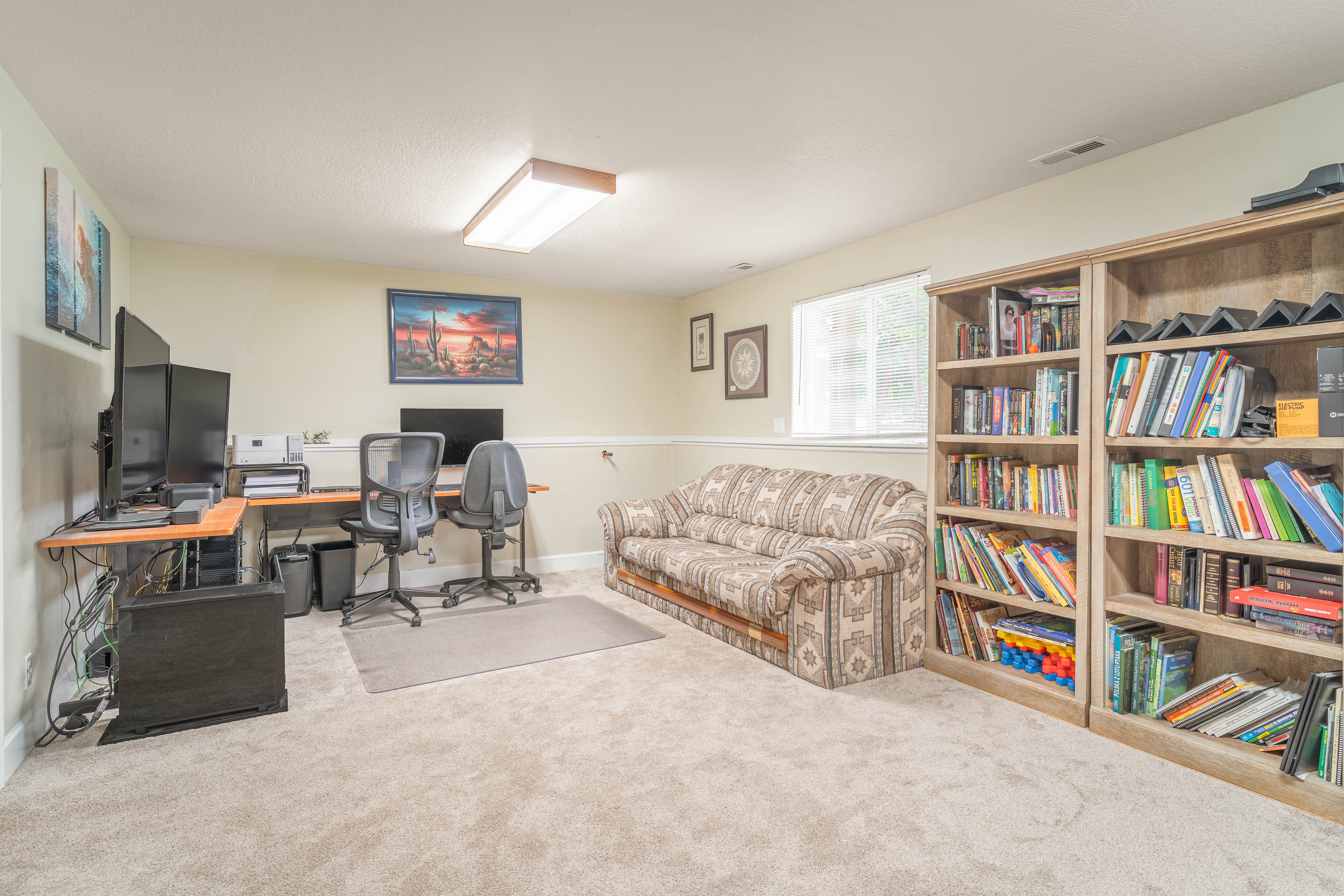 Carpeted room featuring a window, built-in shelving, an L-shaped desk, and a drop ceiling with fluorescent lighting