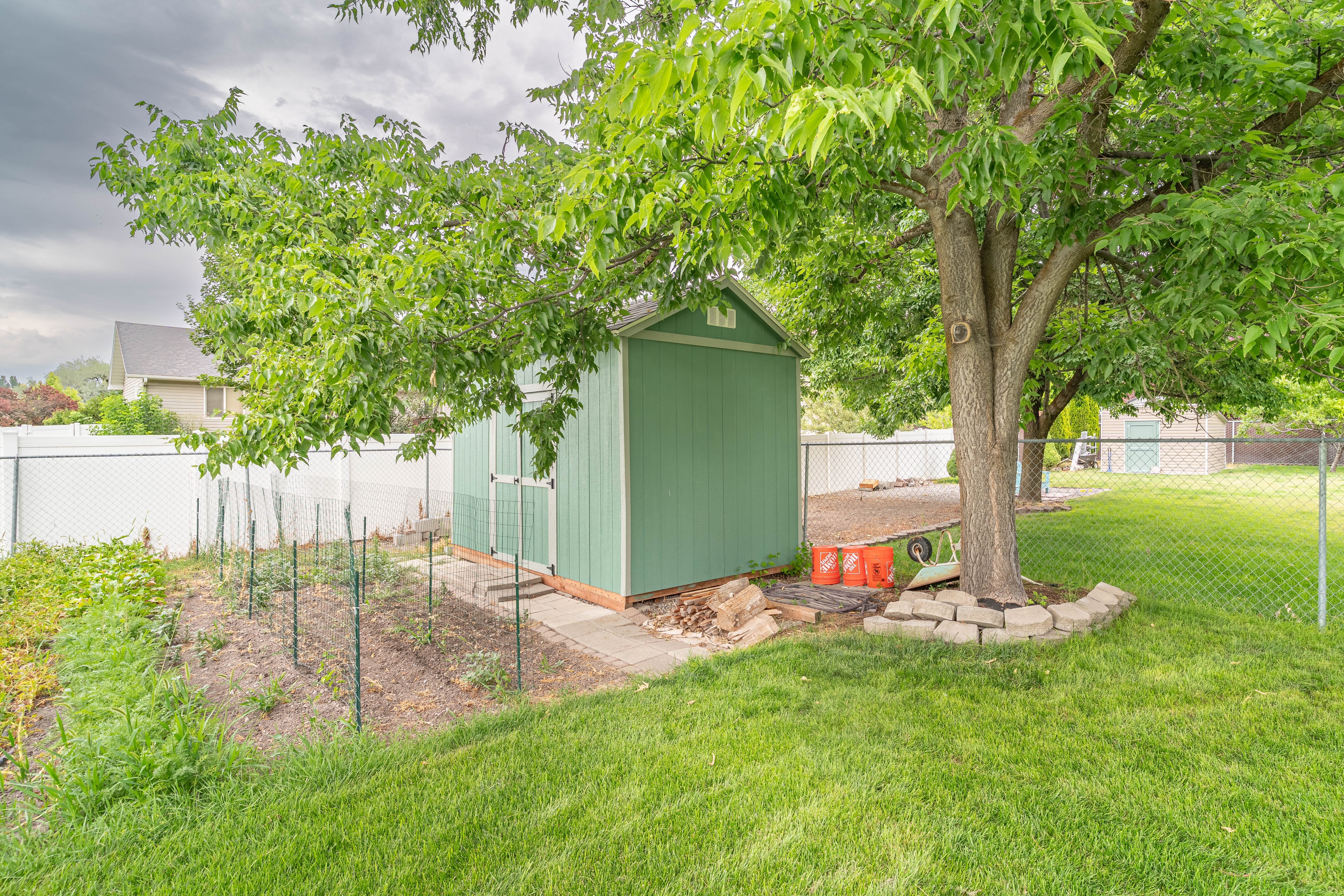 Spacious backyard featuring a green storage shed, mature tree with paver stone base, chain link fencing, and a cultivated garden area