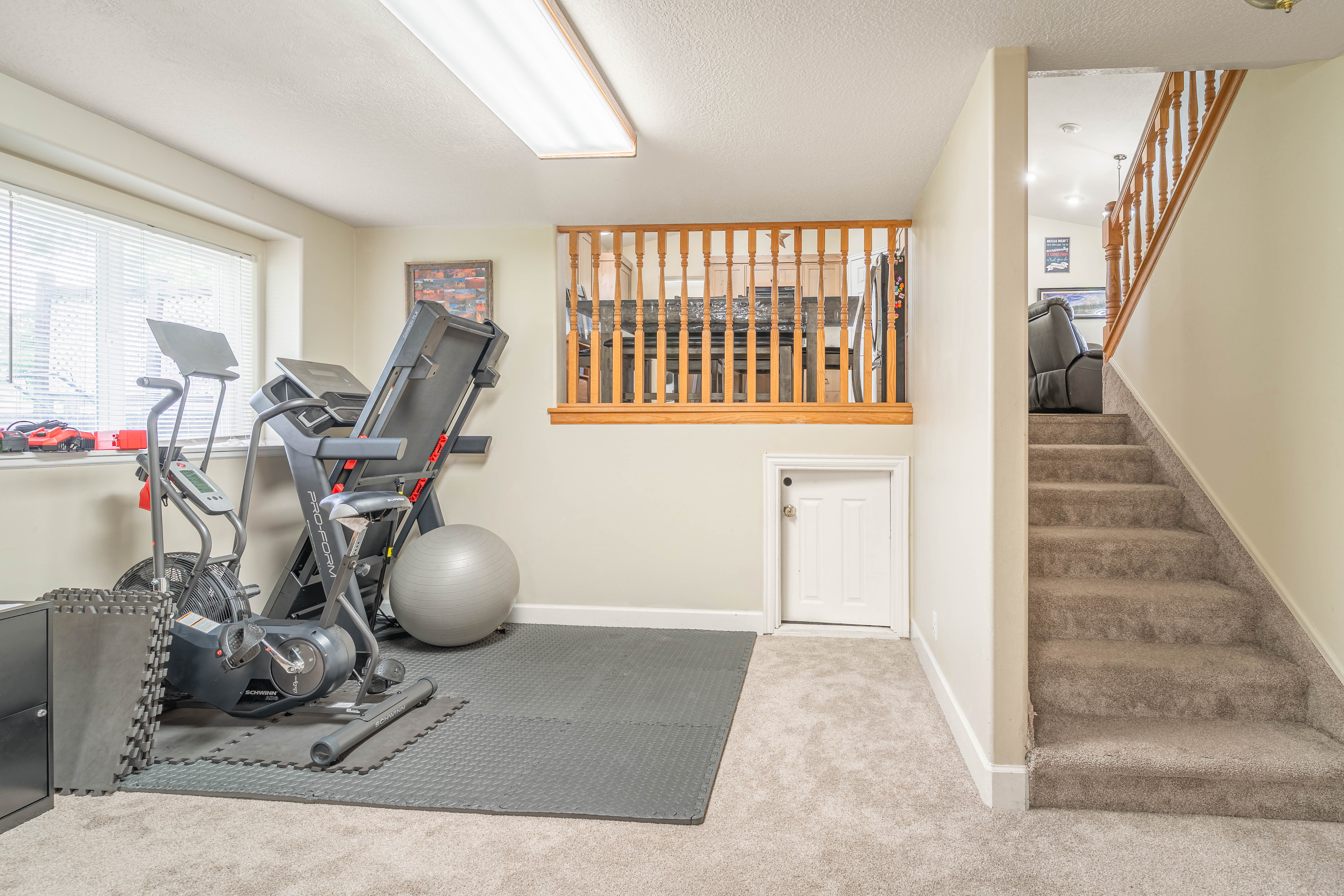 Carpeted lower level featuring a window with blinds, a small white door, a carpeted staircase with wood railing, and a half-wall with wood railing