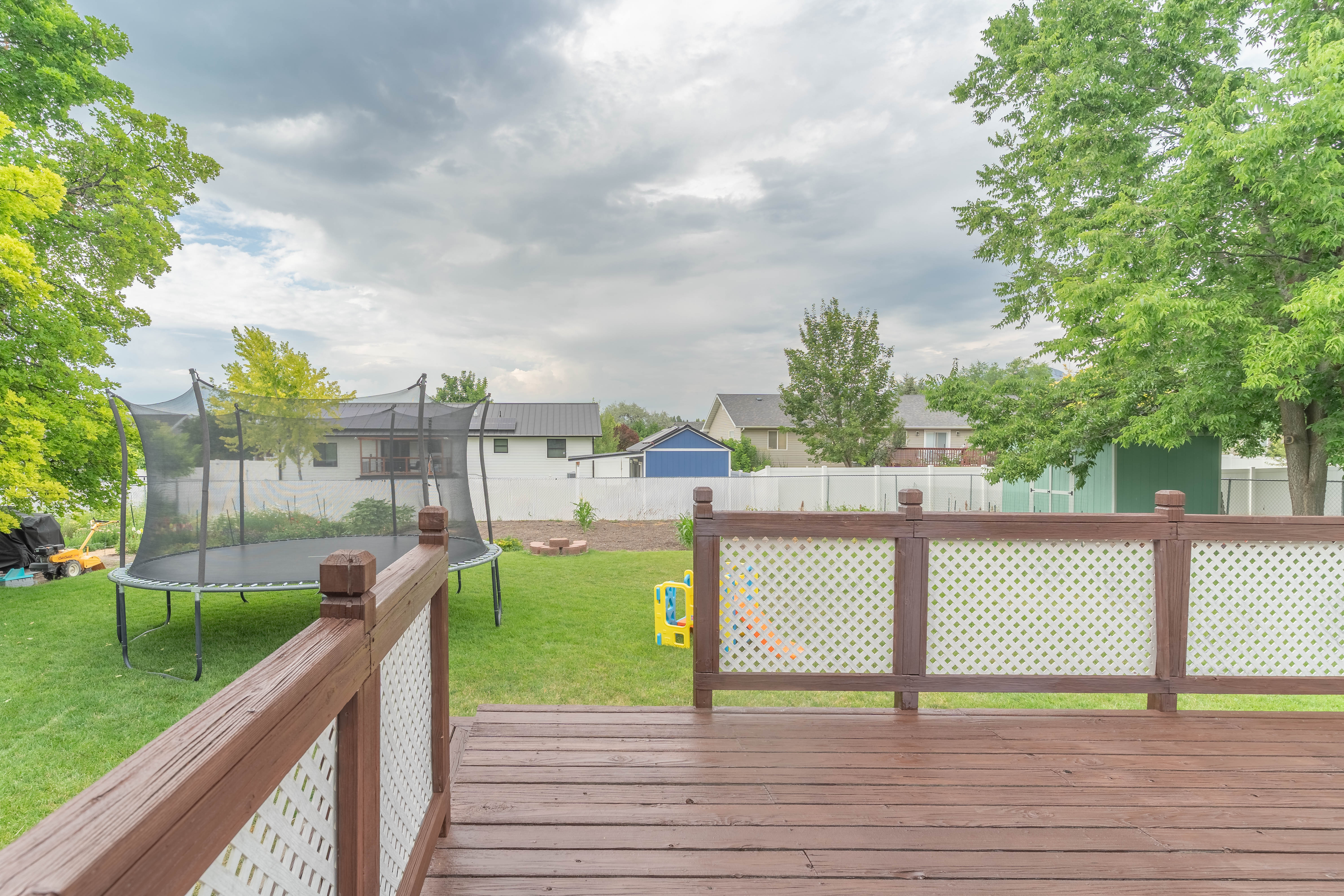 Spacious backyard featuring a wood-finish deck with lattice railing, mature trees, and a green storage shed