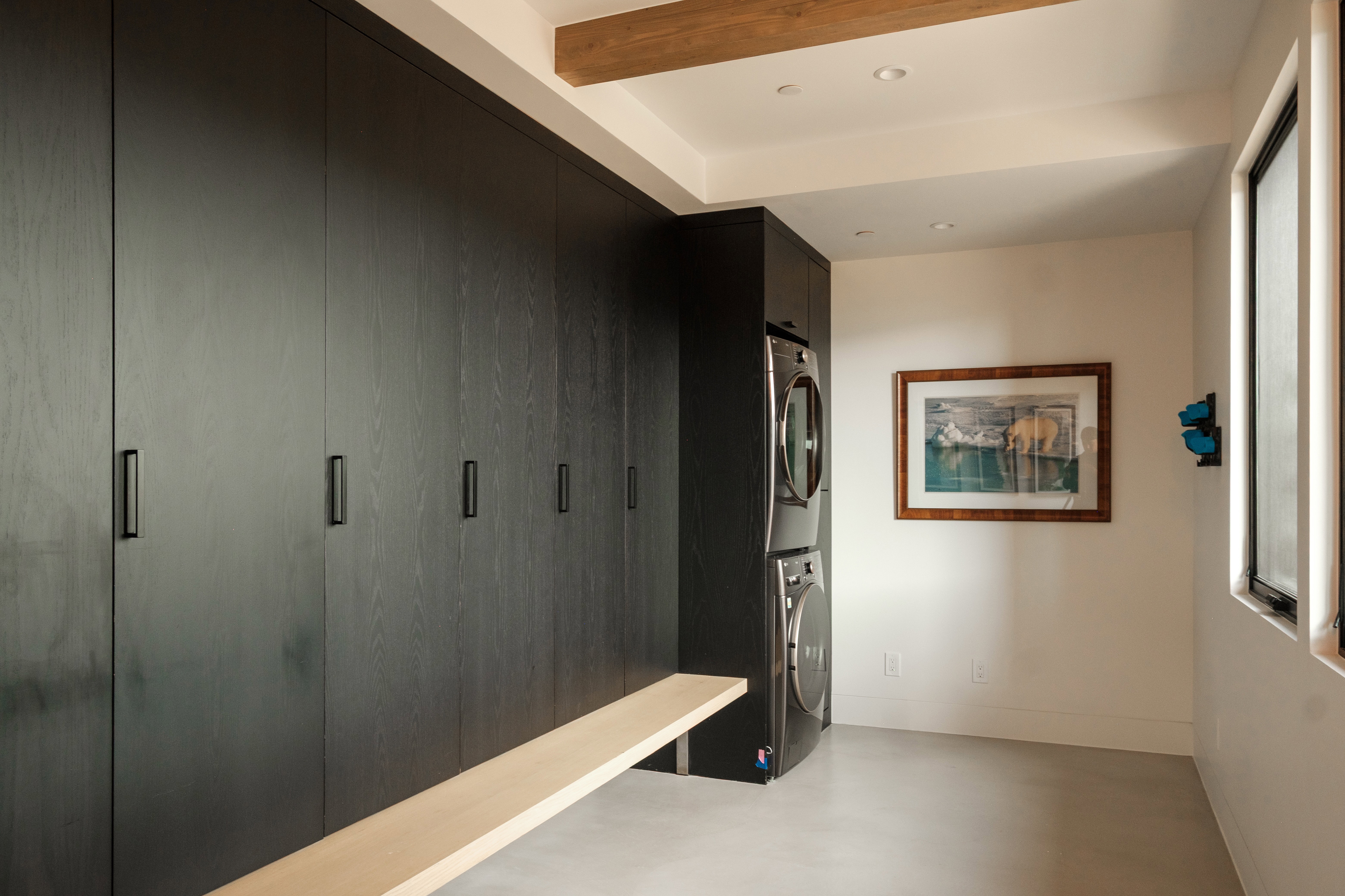 Full-height dark wood-finish cabinetry with modern black pulls, integrated stacked washer and dryer, and a natural wood bench